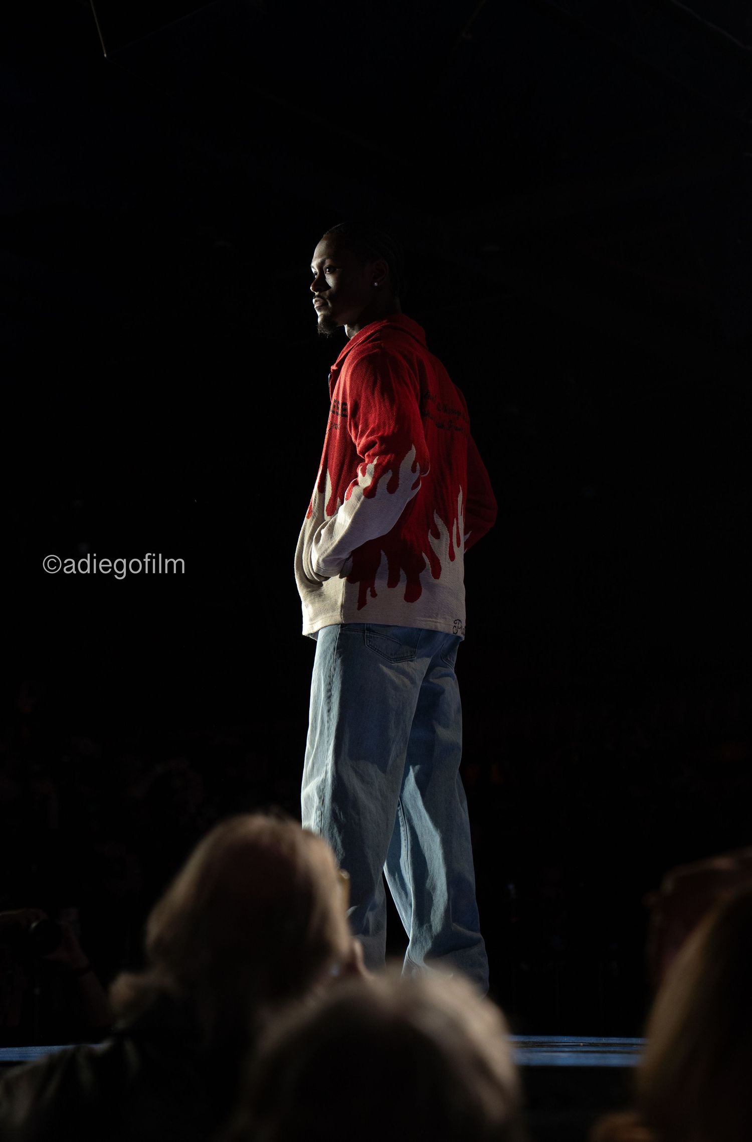 A performer in a red and white jacket stands on a dark stage with audience silhouettes visible below.