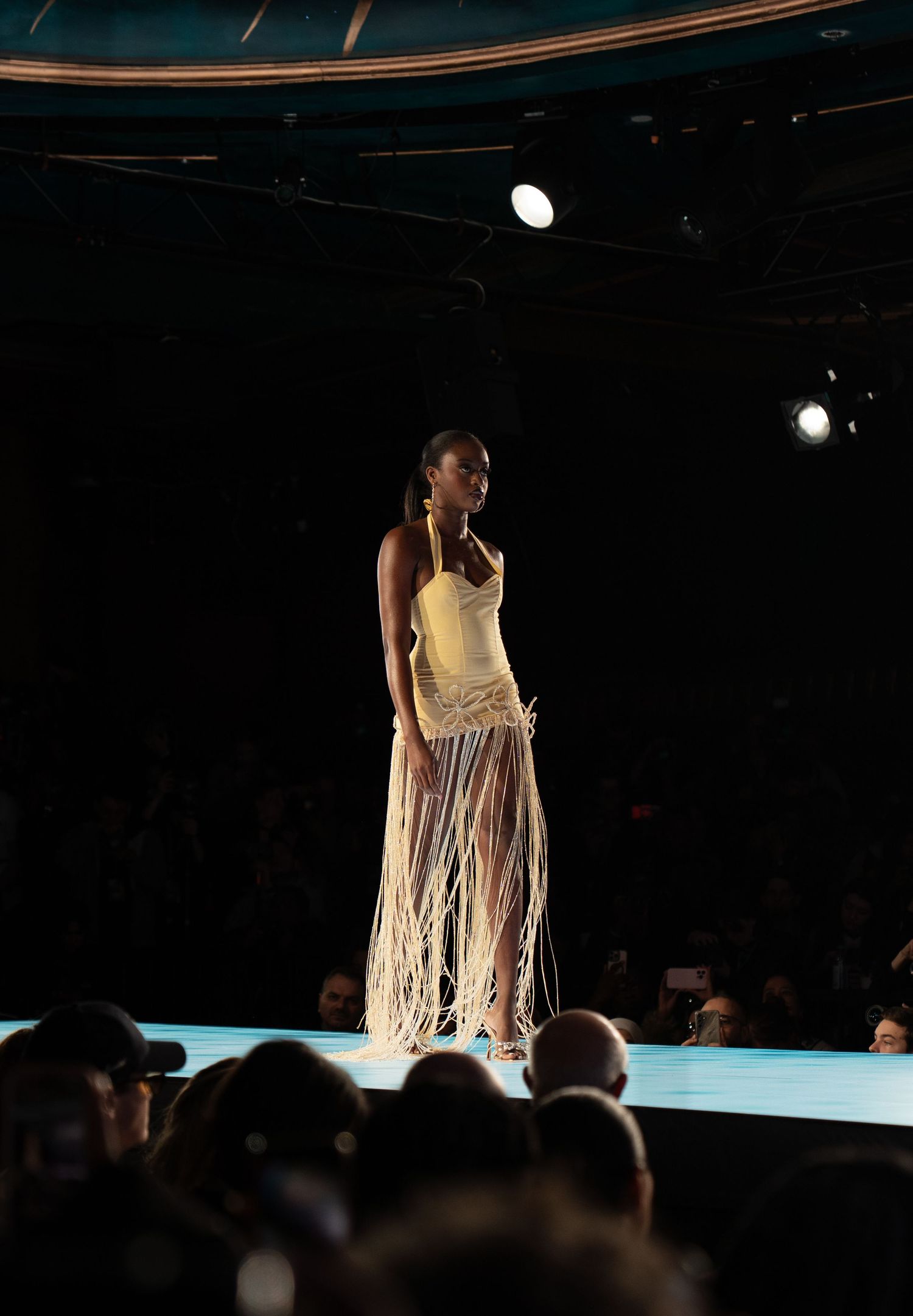 Models showcase elegant evening wear on a dimly lit runway with dramatic spotlighting during a fashion show.