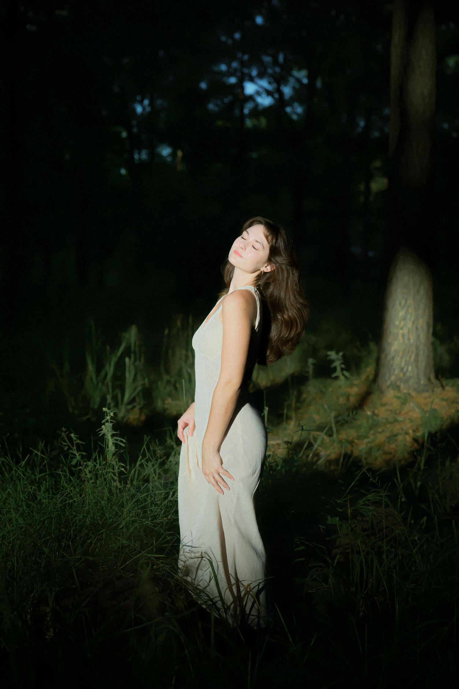 Artistic forest portrait with figure in flowing white dress against shadowy trees.