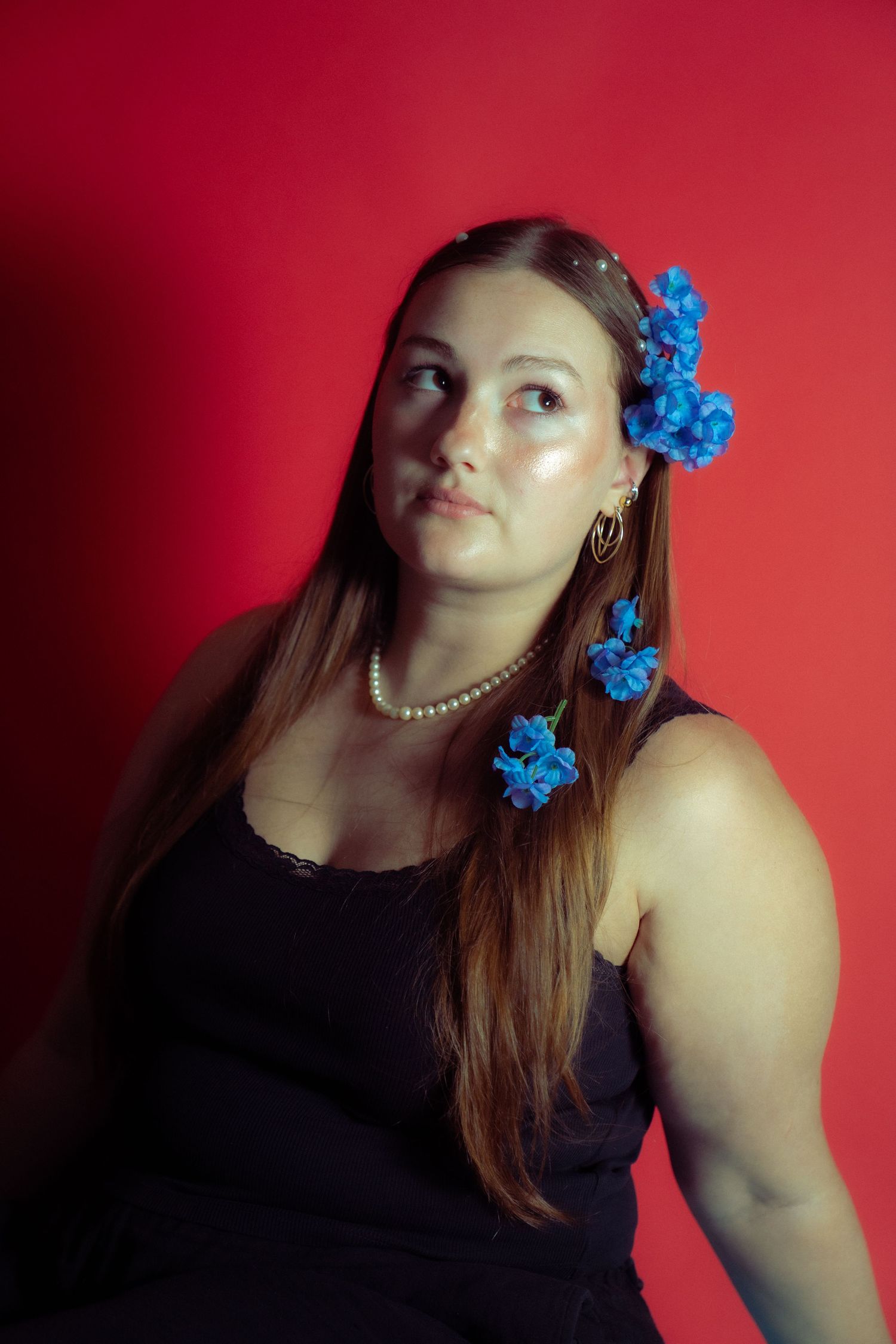 Portrait against a red backdrop with long brown hair decorated with bright blue flowers and a pearl necklace.