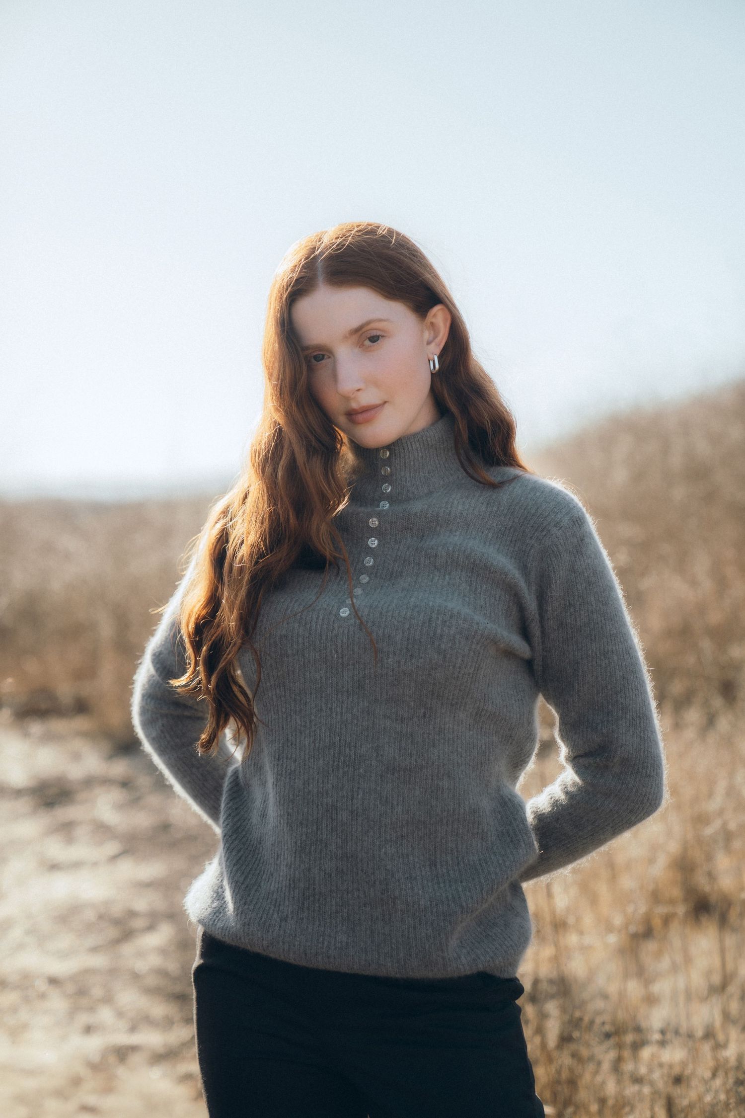 Person in a gray turtleneck sweater poses in a field during golden hour with warm natural lighting.