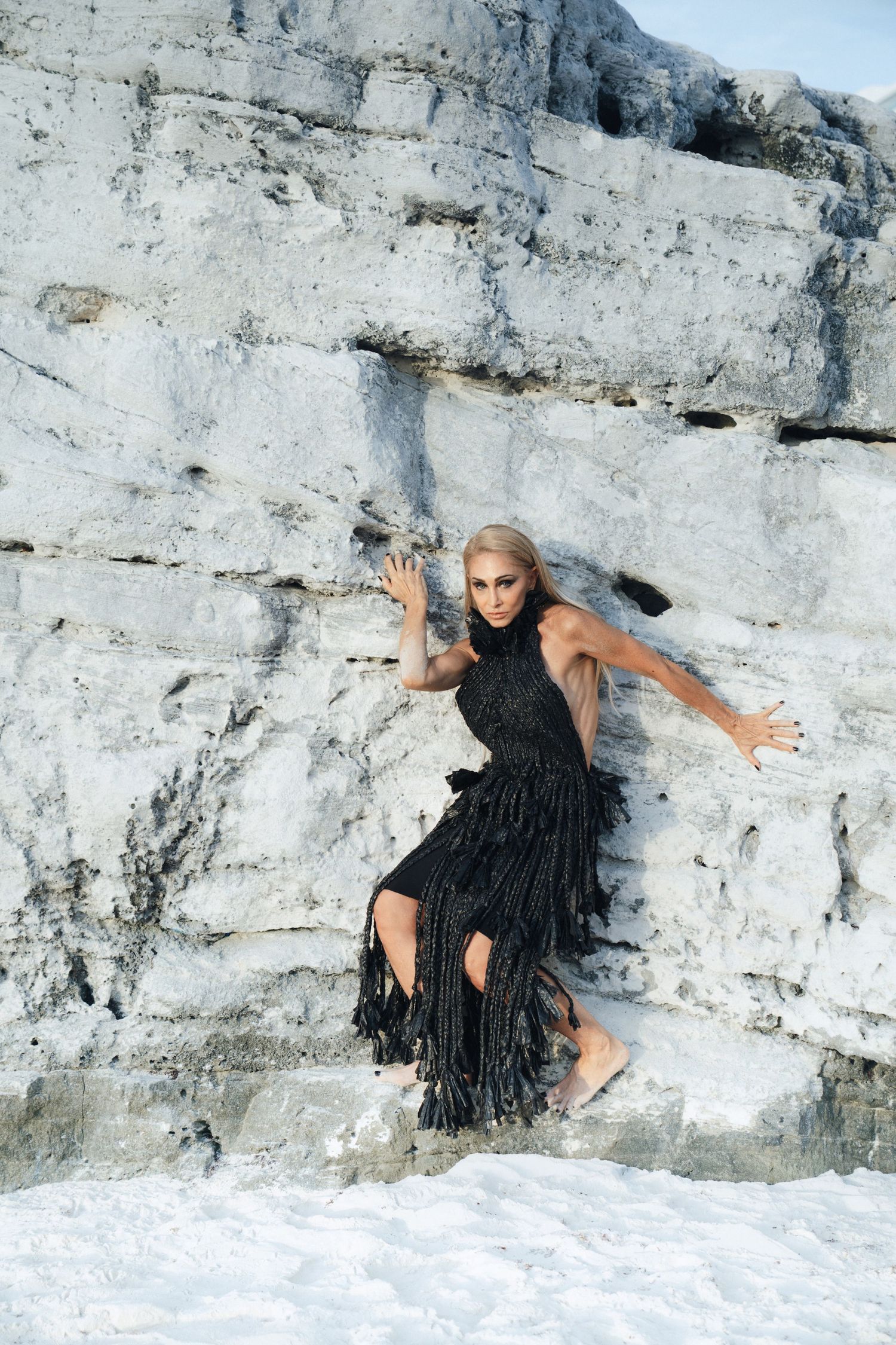 A figure in a black dress poses dramatically against a white limestone rock wall in an artistic photo series.