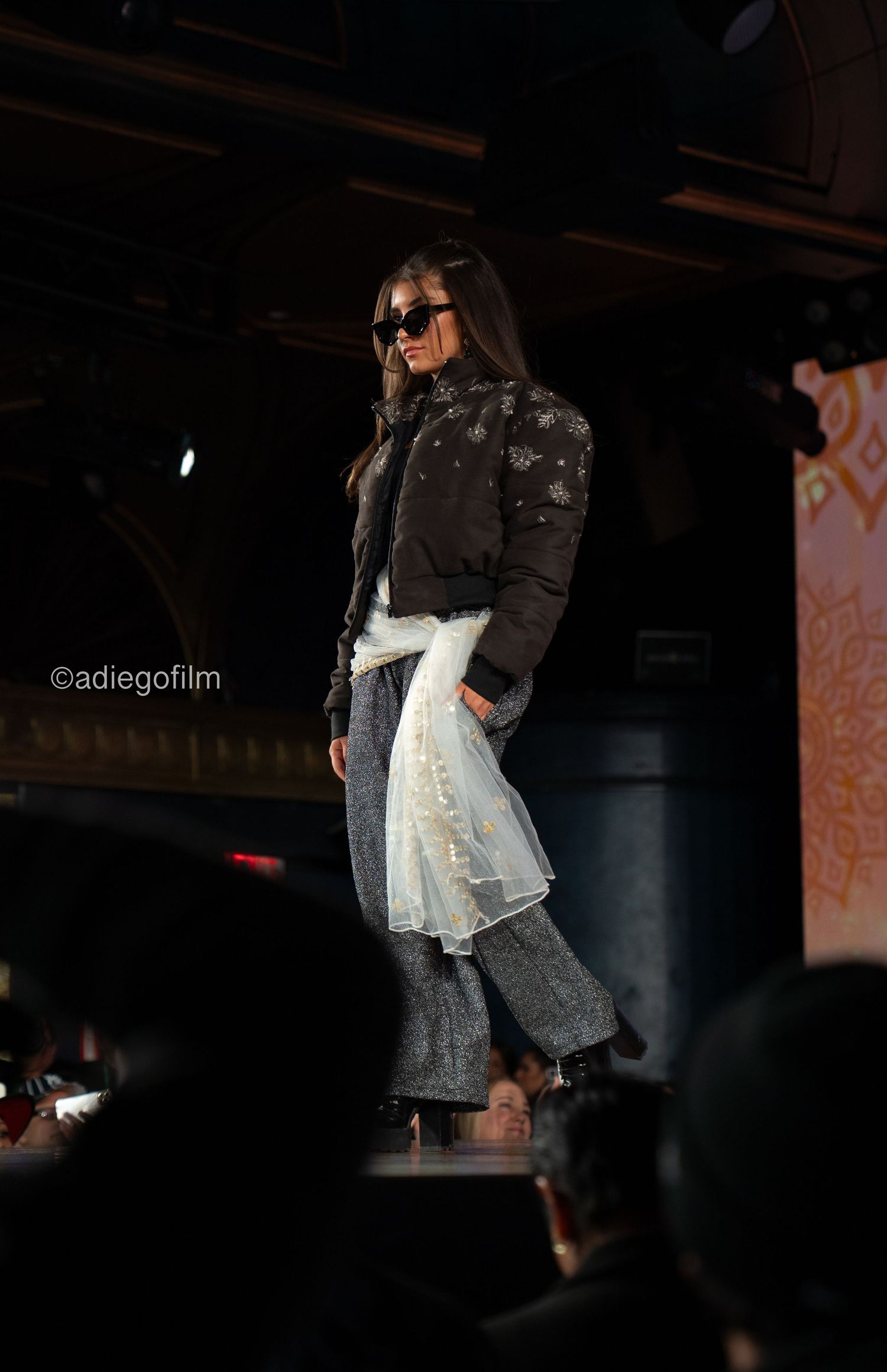 Model walks runway in edgy black studded jacket with white layered skirt and wide-leg jeans during fashion show.