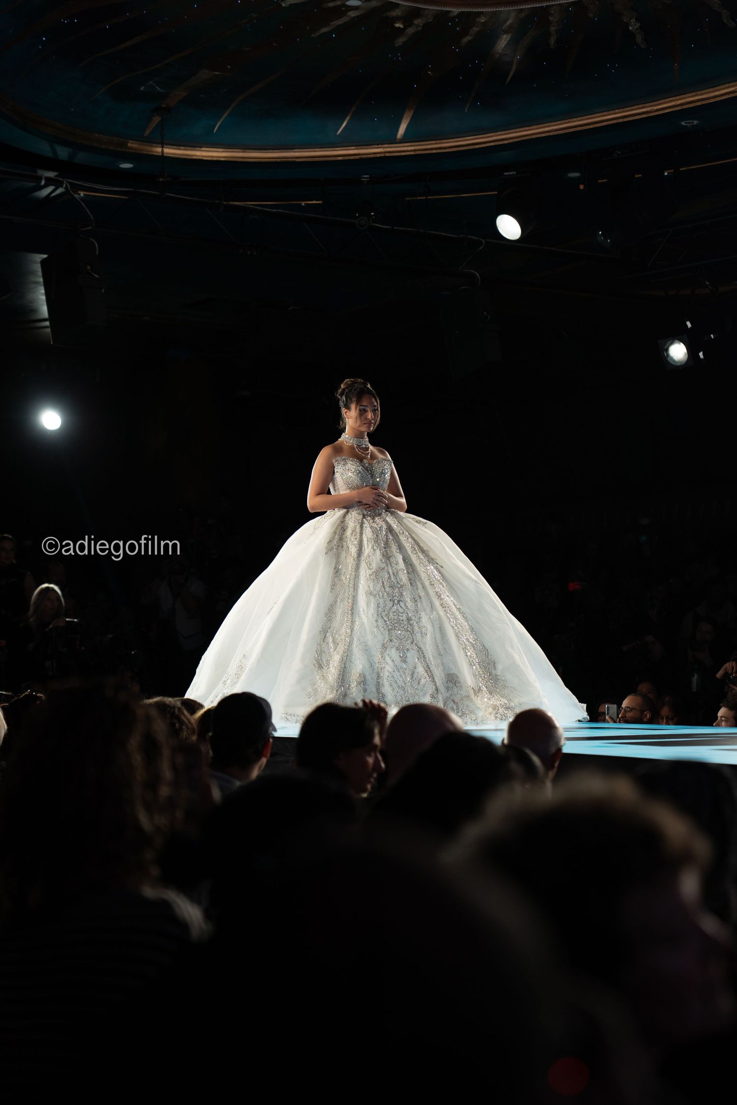 A model walks the runway in a dramatic white ball gown with a voluminous skirt at a fashion show.