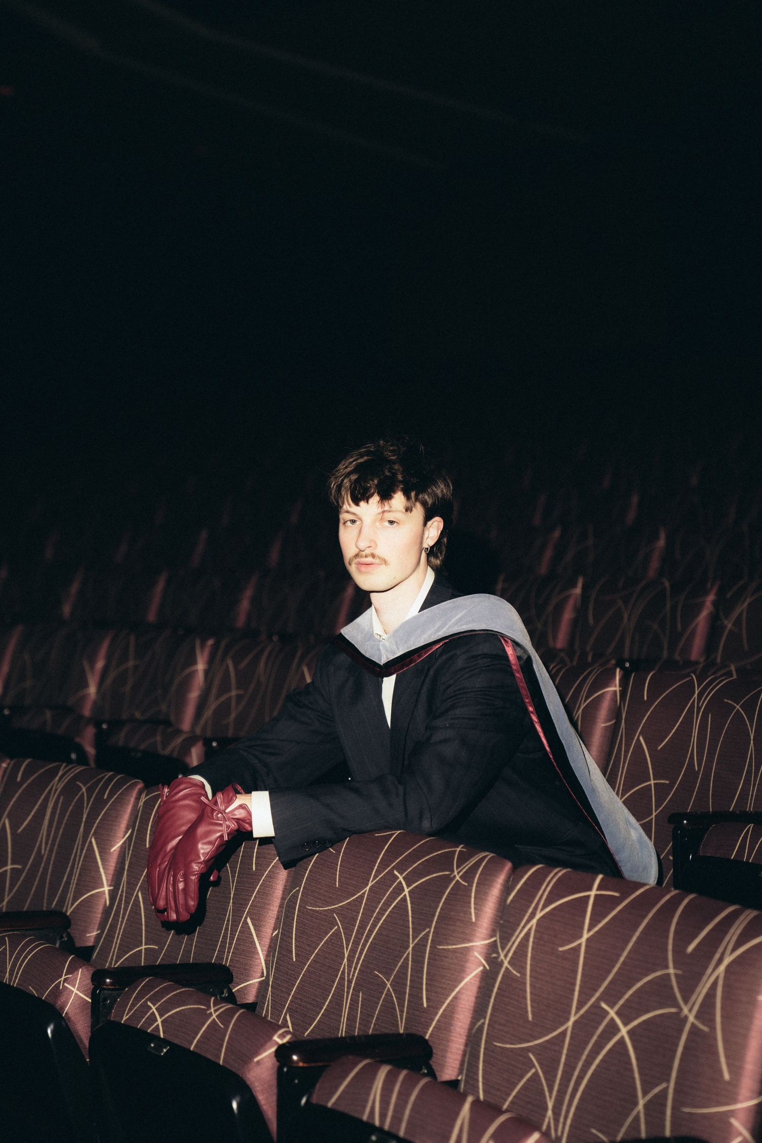 A person in a suit sits in a dark theater with rows of empty seats visible in the background.