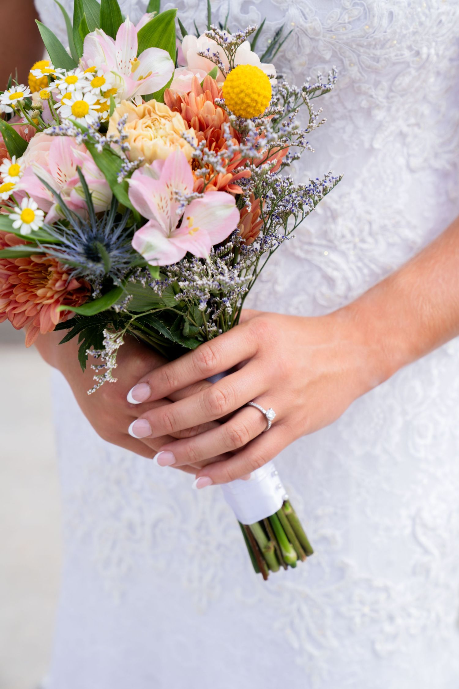 A bridal bouquet featuring pink, yellow and orange flowers with delicate daisies and a wedding ring.