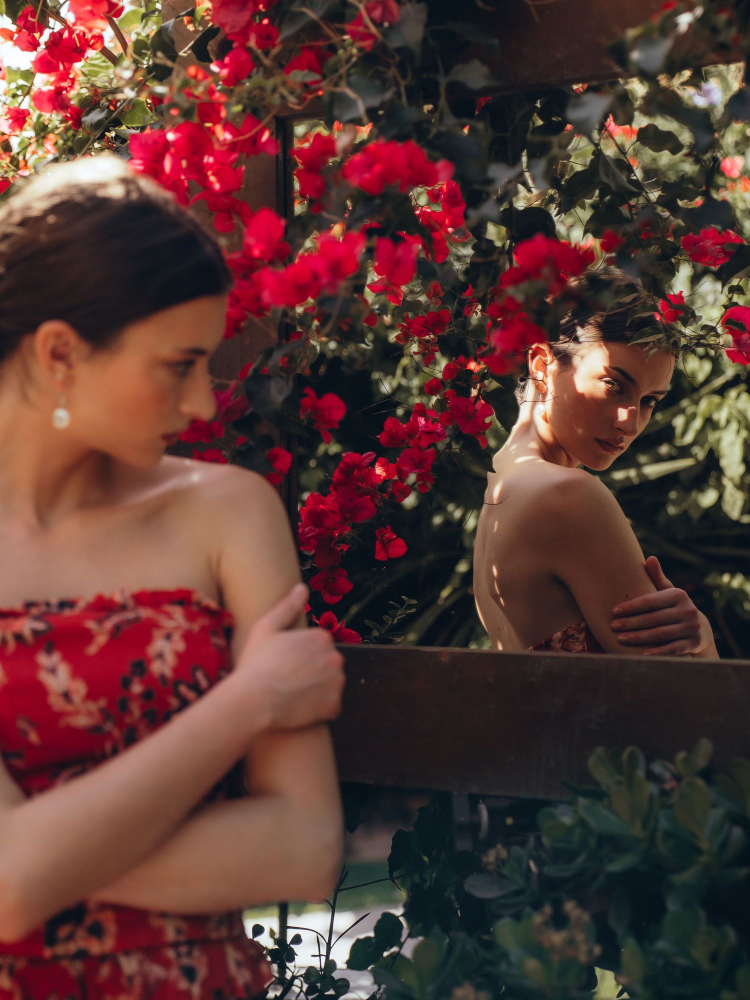 Two figures pose in a romantic garden setting with vibrant red bougainvillea flowers creating a dramatic backdrop.