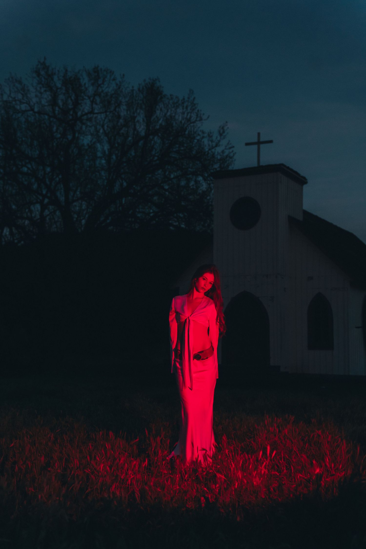 Glowing red figure stands eerily in a dark field against a moody nighttime backdrop with a church silhouette.