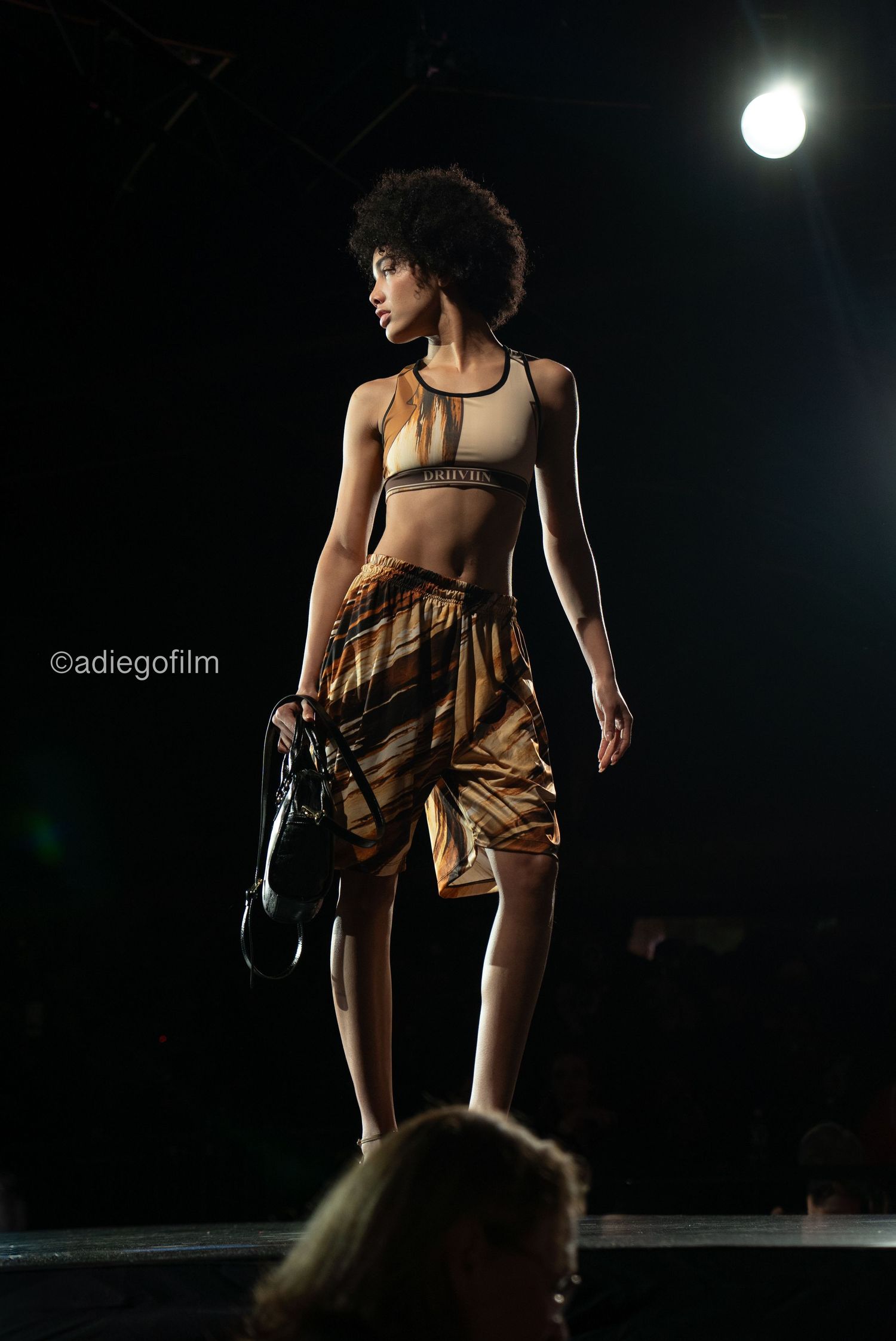 Models showcase casual summer outfits on a dimly lit catwalk with dramatic spotlighting.