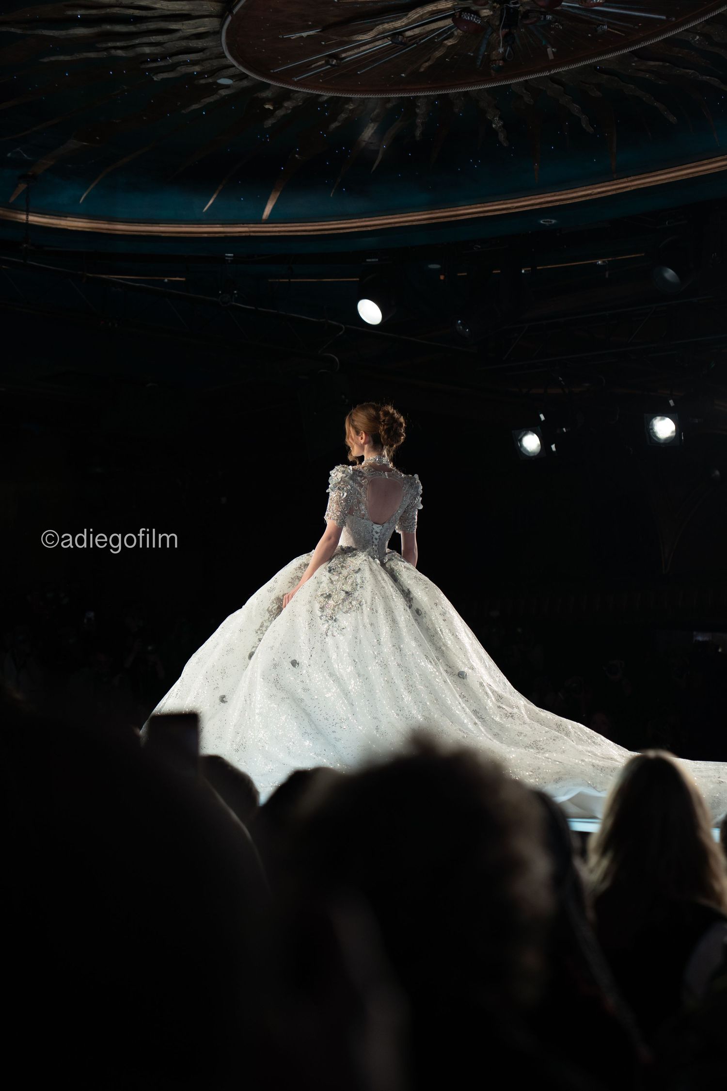 A bride in a flowing white wedding gown twirls on a dark stage with dramatic spotlighting creating an ethereal effect.