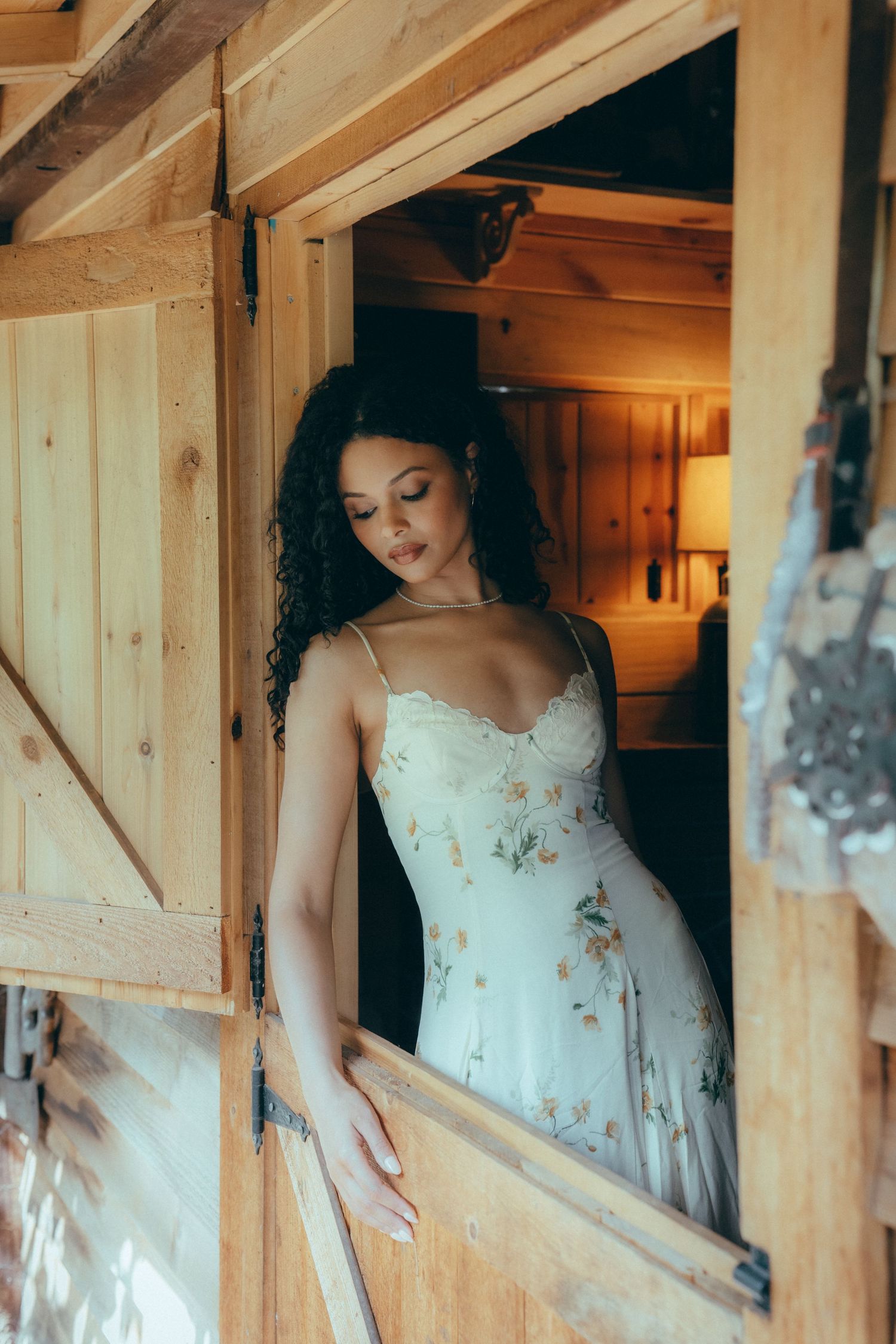 Woman in floral sundress standing in rustic wooden barn doorway with warm interior lighting.