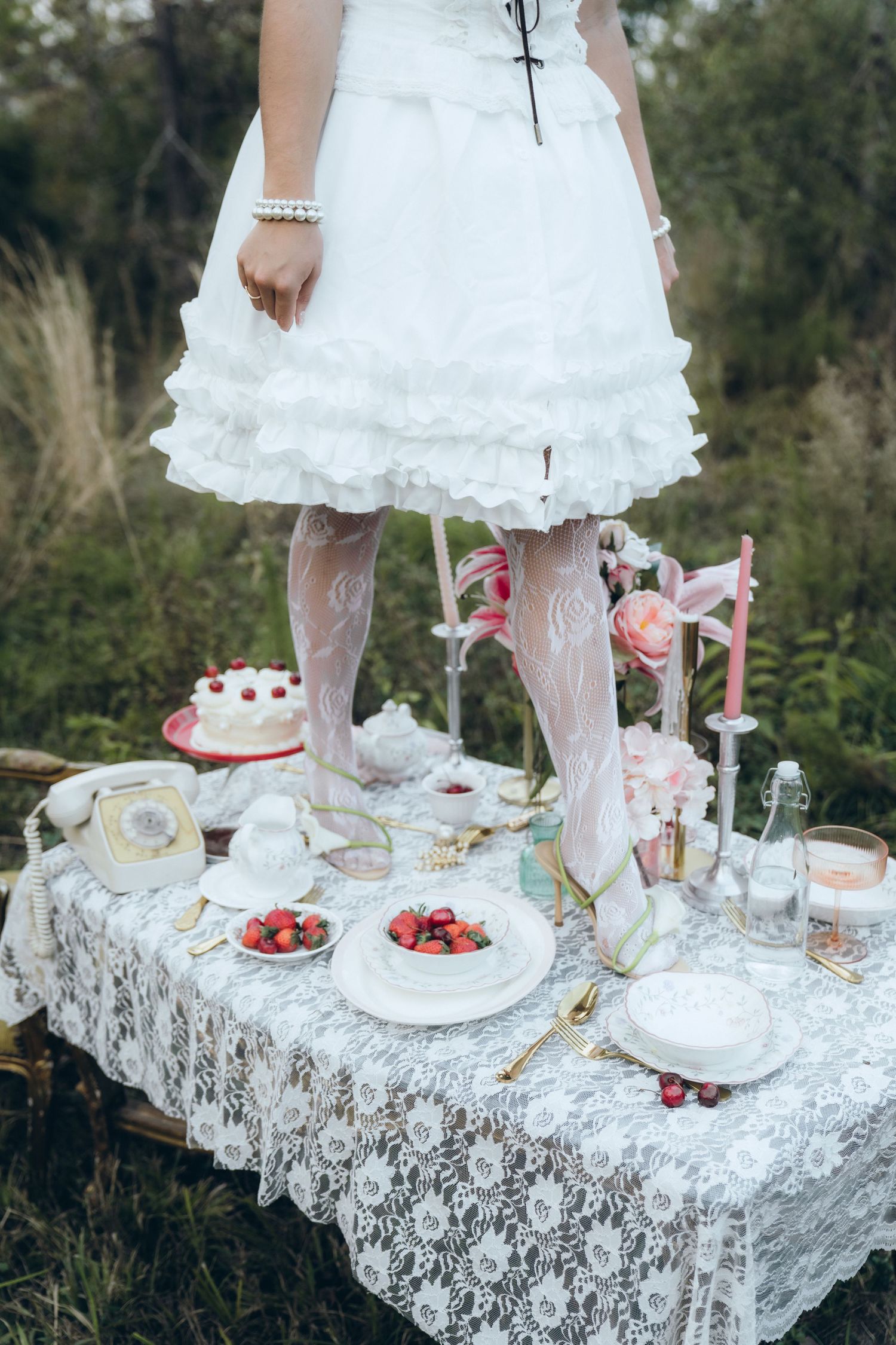 A whimsical tea party setup with white lace tablecloth, strawberry desserts, and someone in a white dress and lace stockings.