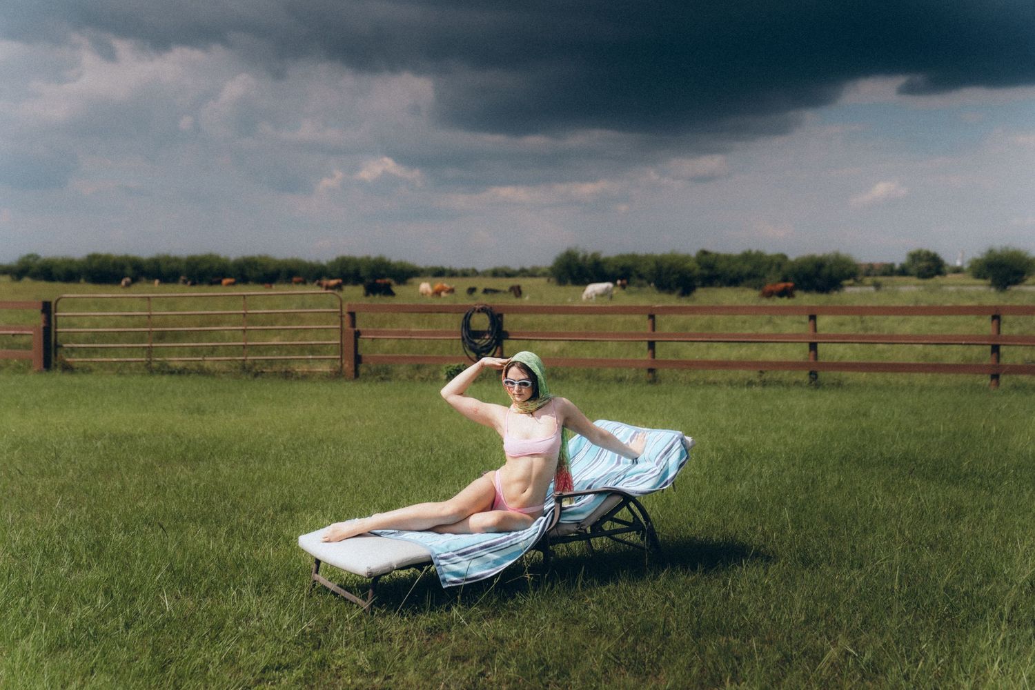 A person relaxes on a lounge chair in a sunny green pasture with a wooden fence and stormy clouds overhead.