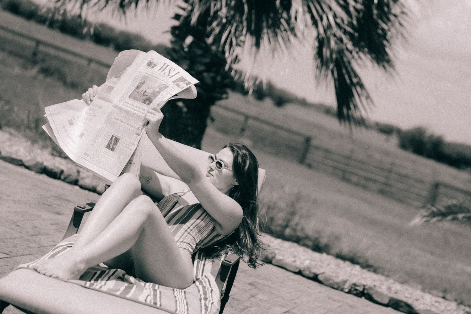 Young woman in a swimsuit lying on a blanket reading a newspaper outdoors on a sunny day in black and white.