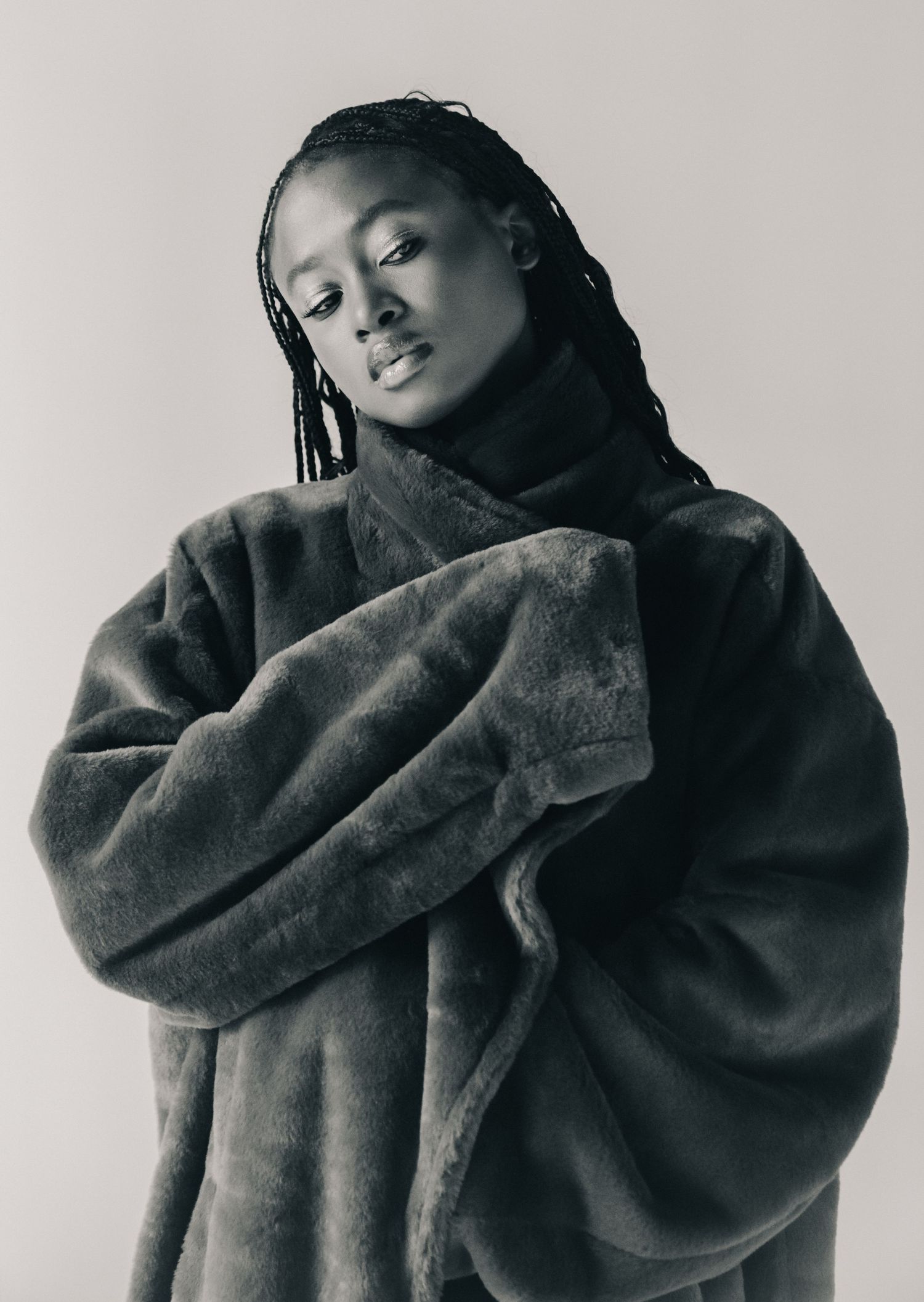 Black and white portrait of a model in an oversized faux fur coat with braided hair against neutral backdrop.