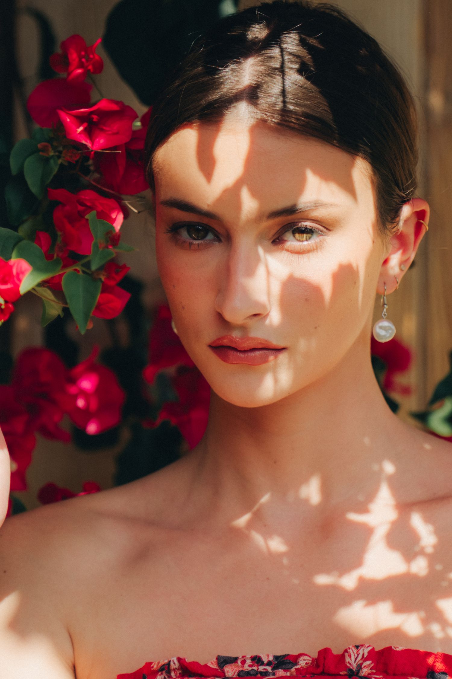 Young woman with pearl earring surrounded by red bougainvillea flowers, dappled sunlight casting shadows on her face.