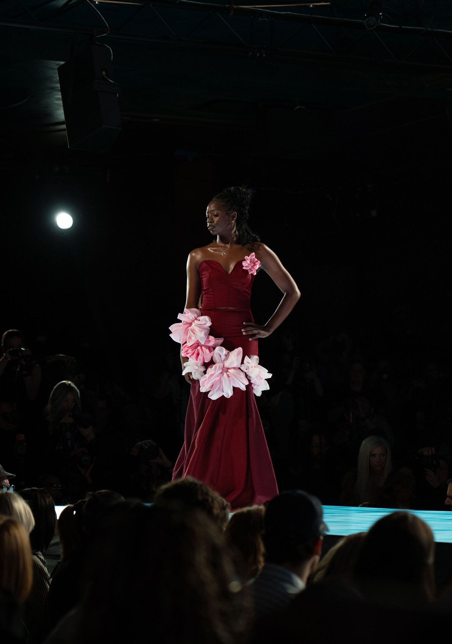 Model in a red strapless dress with pink floral accents walks down a dark runway during a fashion show.