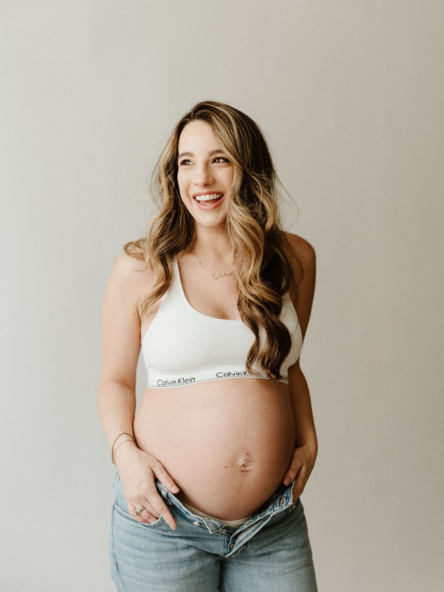 Pregnant woman in Calvin Klein sports bra and jeans poses against neutral background with natural wavy hair.