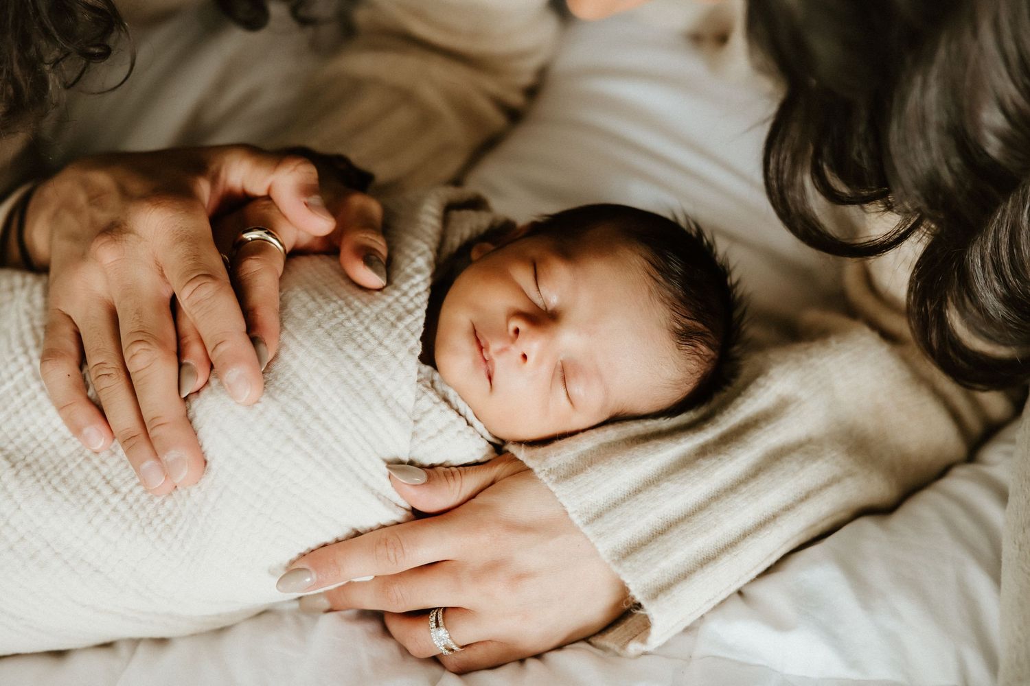 A baby sleeps peacefully in a white bed while a hand gently rests nearby.
