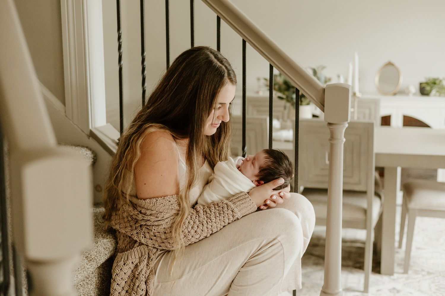 Mother in knit sweater lovingly holding newborn baby by staircase.