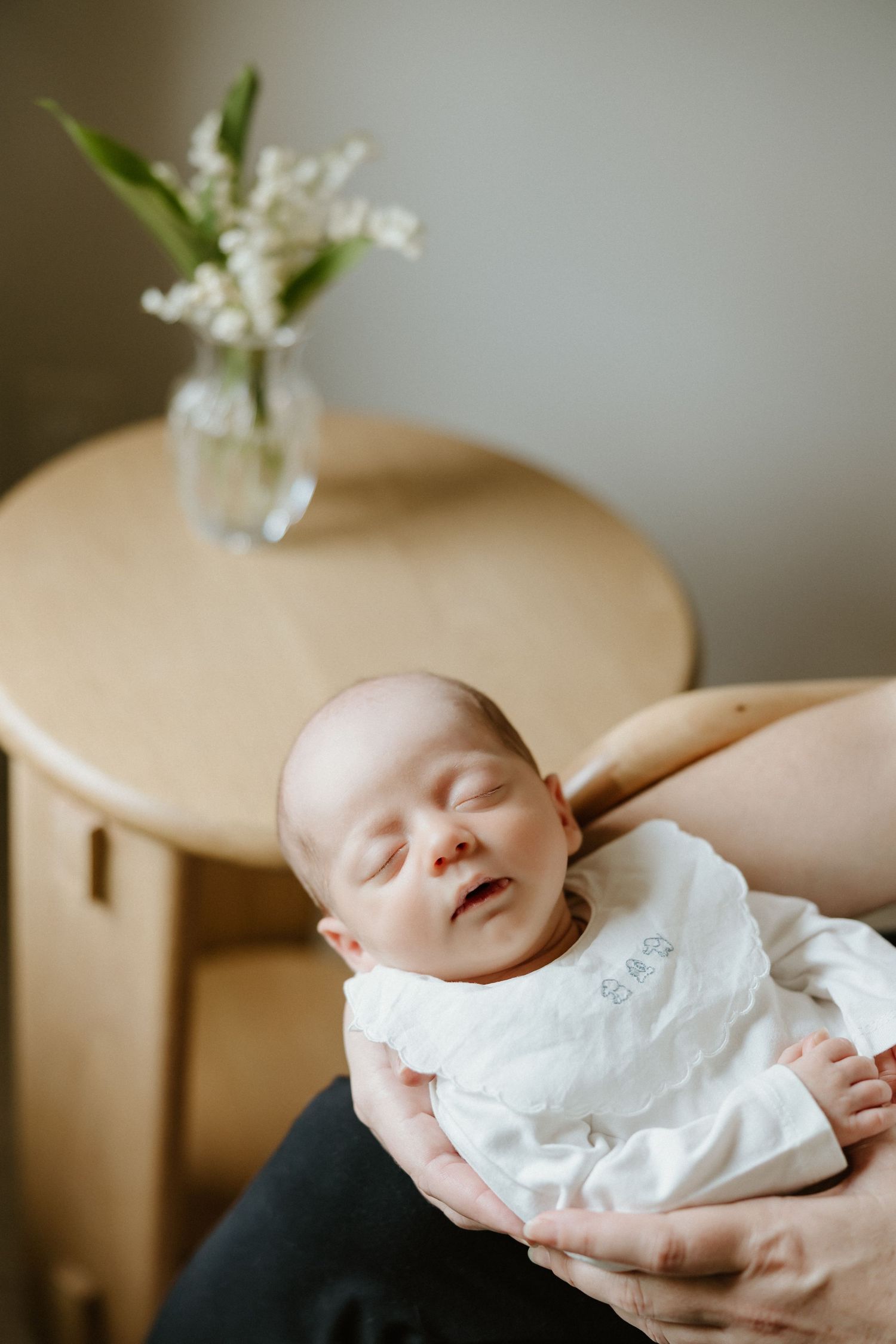 Newborn baby sleeping peacefully in arms, wearing white outfit, with lily of the valley flowers on wooden table behind.