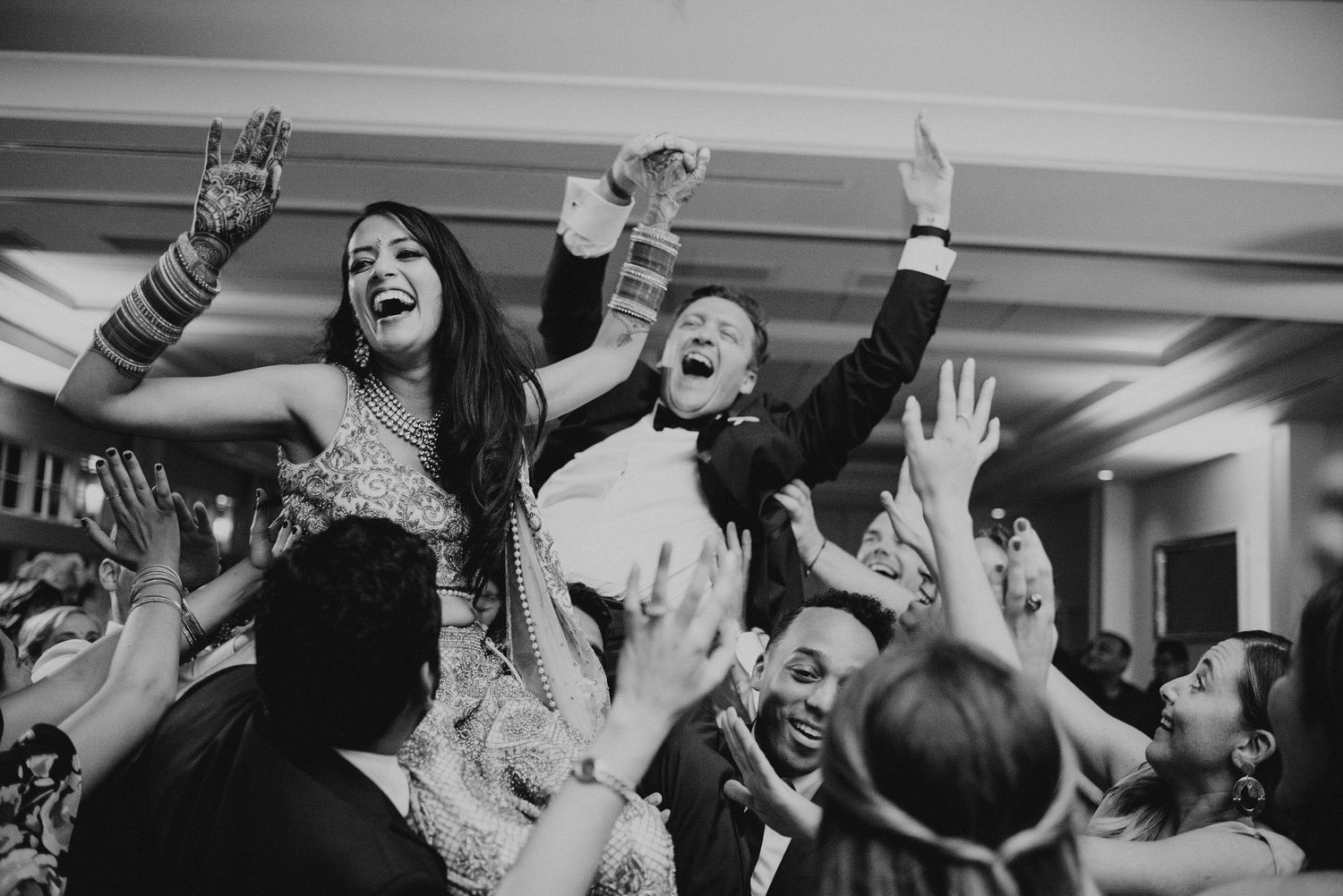 Wedding reception crowd cheering and dancing with arms raised in an indoor venue.