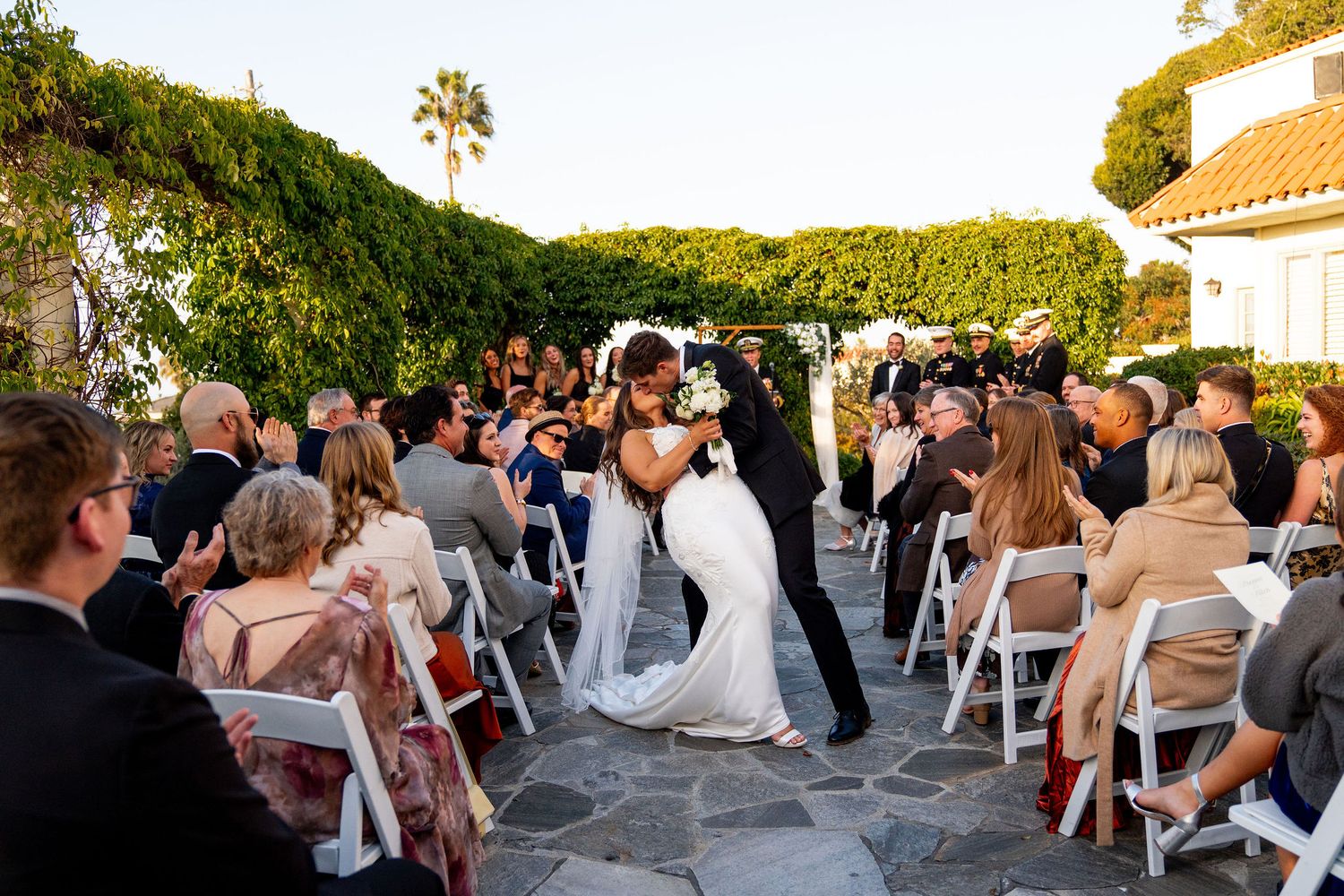 A panoramic view of an outdoor wedding ceremony with guests seated in white chairs at sunset time.