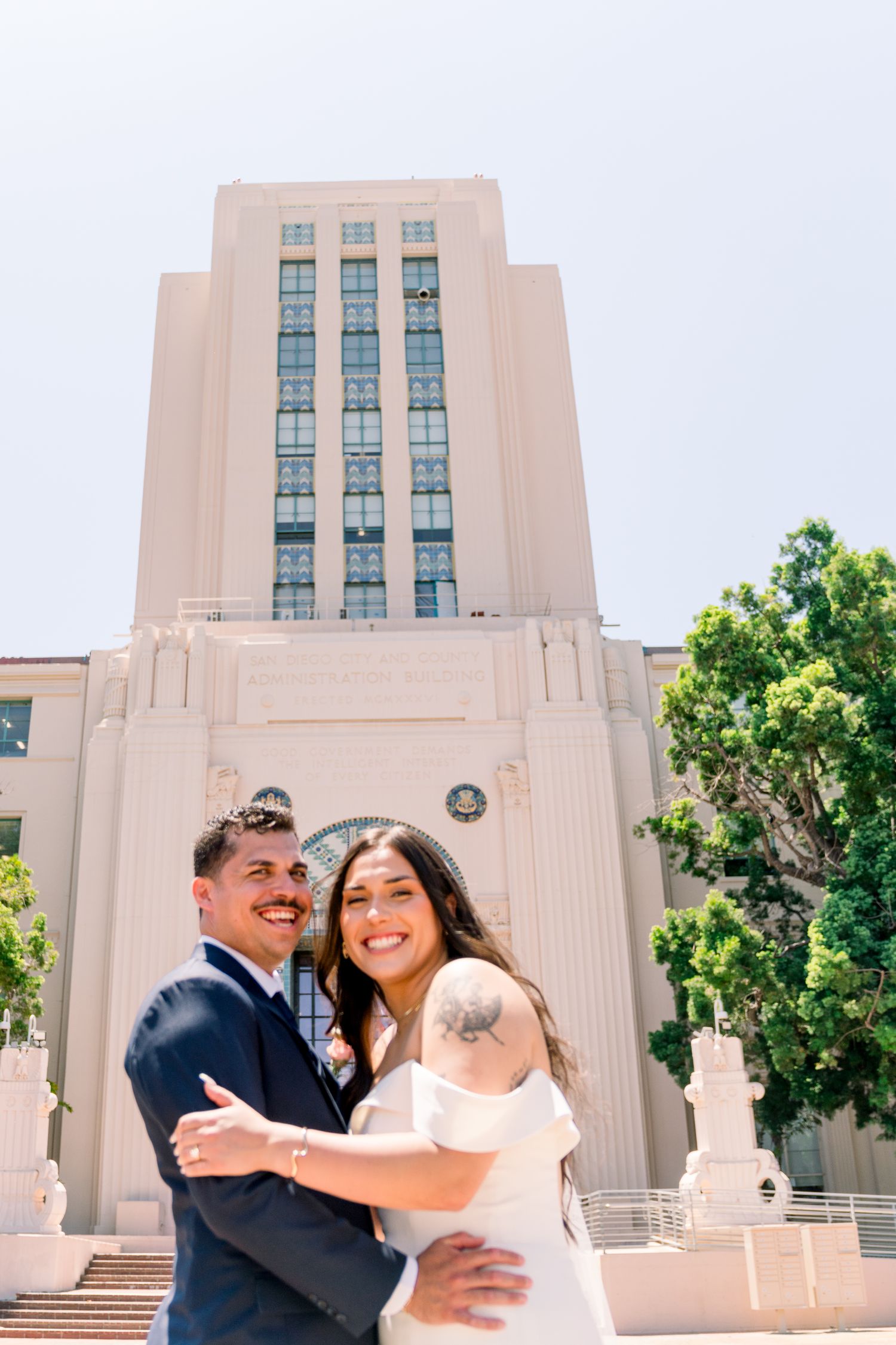 A couple embraces and laughs together in front of an Art Deco style government building on a sunny day.