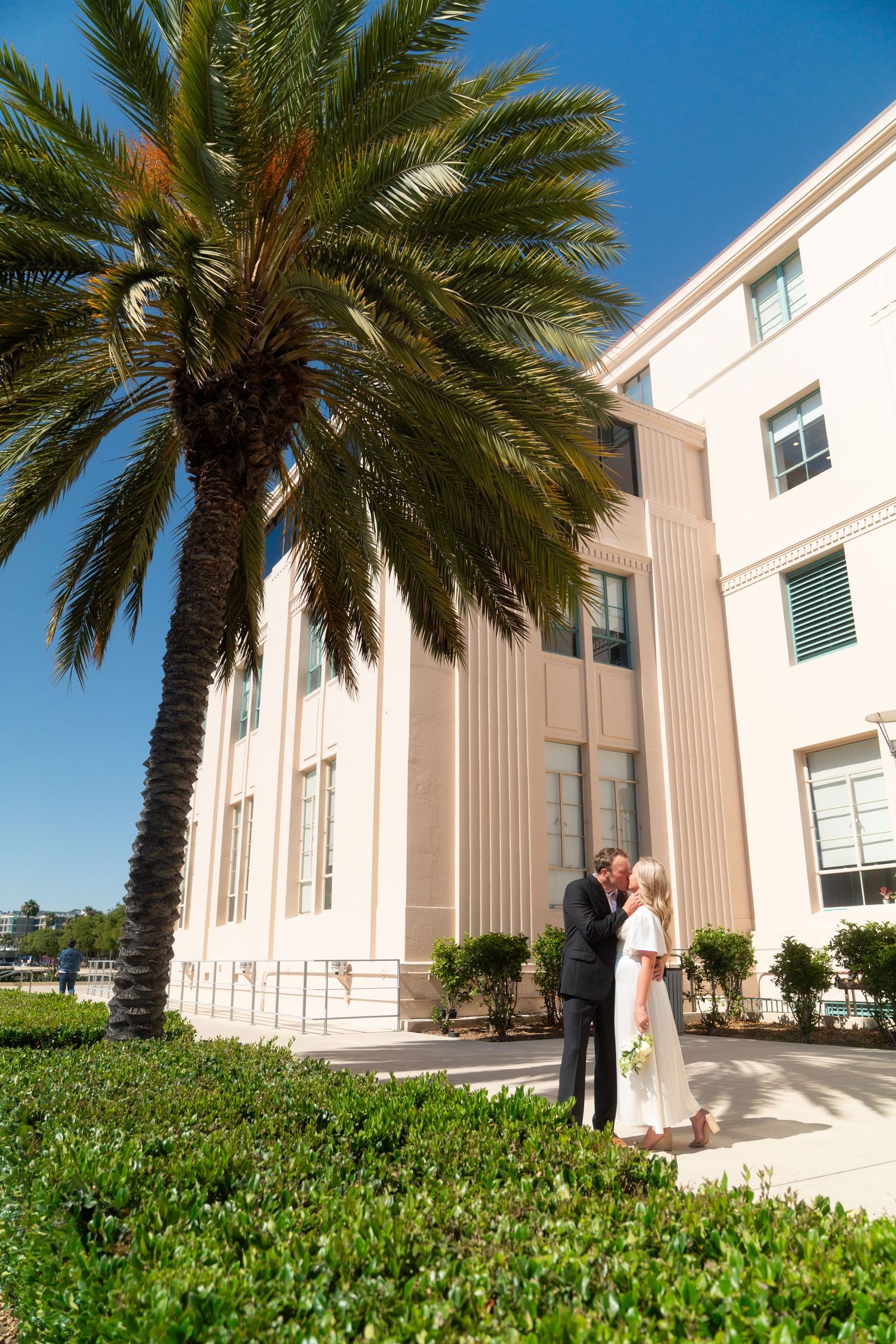Palm trees line the entrance of a modern white building with columns under bright blue sky.