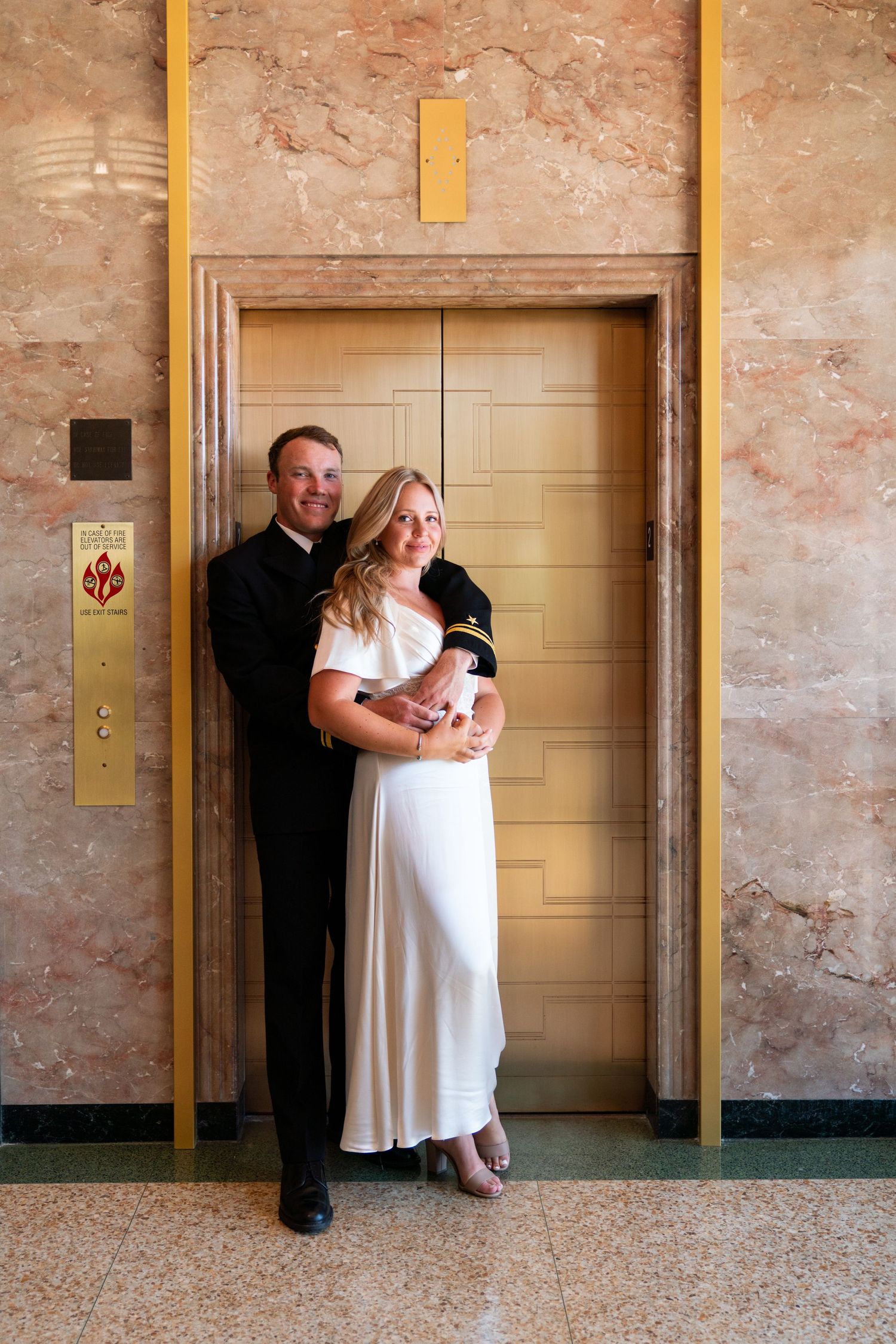 A couple poses together in front of a vintage gold elevator door in an art deco style hallway.