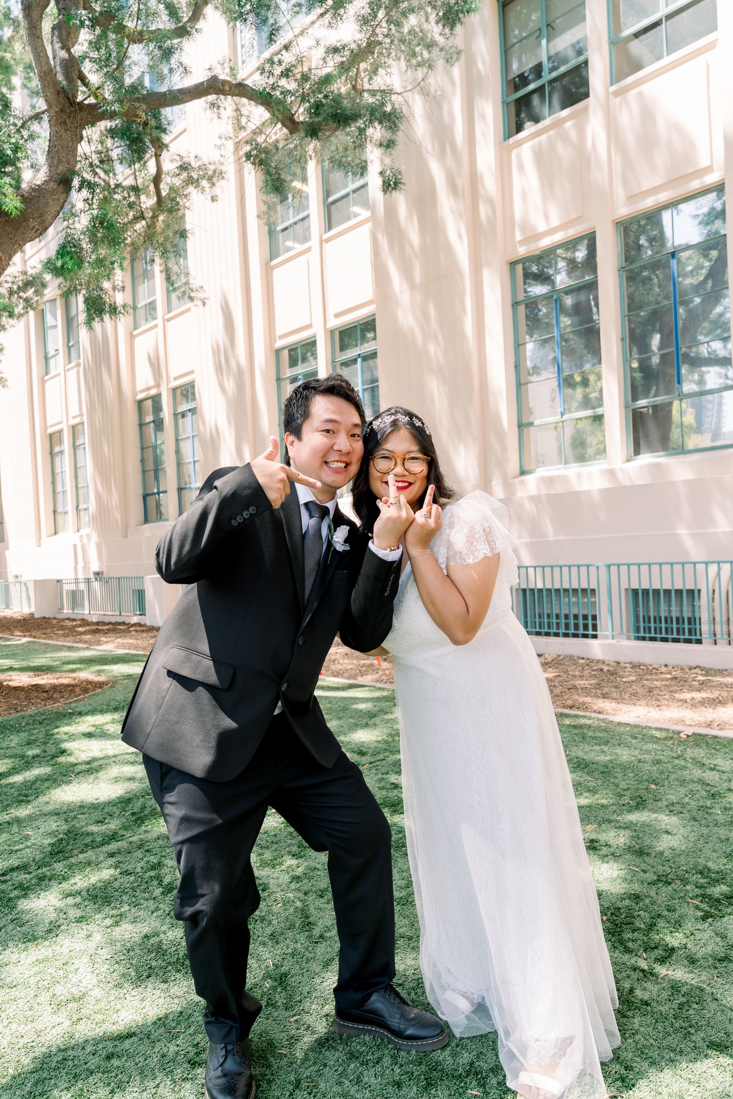 A joyful couple in formal wedding attire pose playfully on a sunlit lawn in front of a light-colored building.