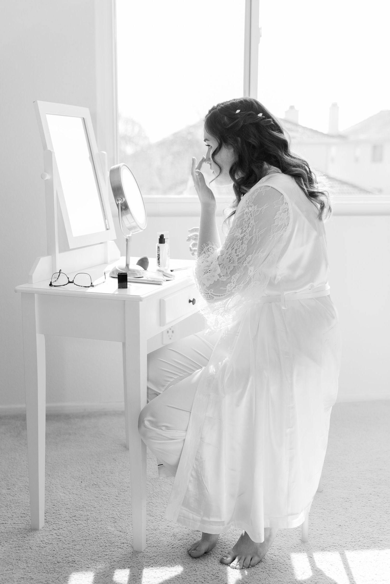 A figure in a flowing white robe sits at a vanity table with a mirror in a sunlit room with soft, dreamy lighting.