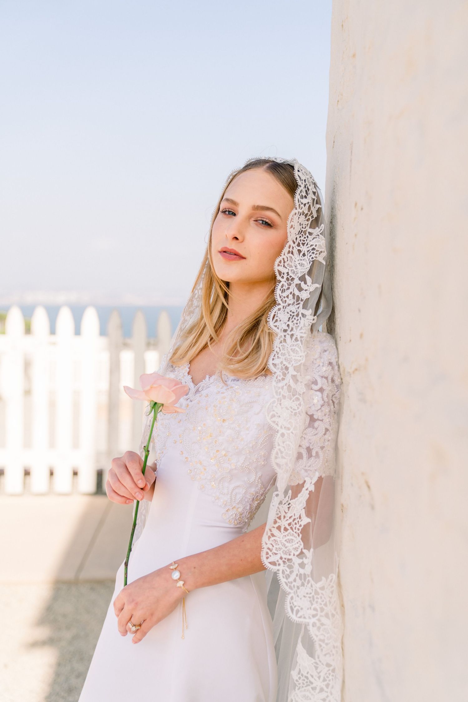 Bride in elegant white lace wedding dress and veil holds single rose while standing against light wall outdoors.