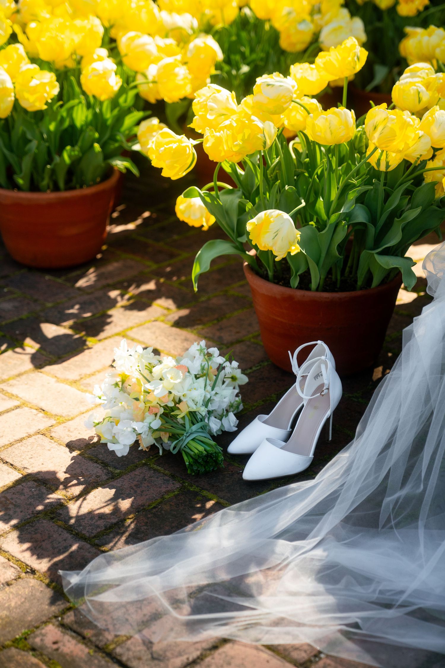 White bridal shoes and veil rest beside potted yellow tulips and white flowers on a sunlit garden patio.