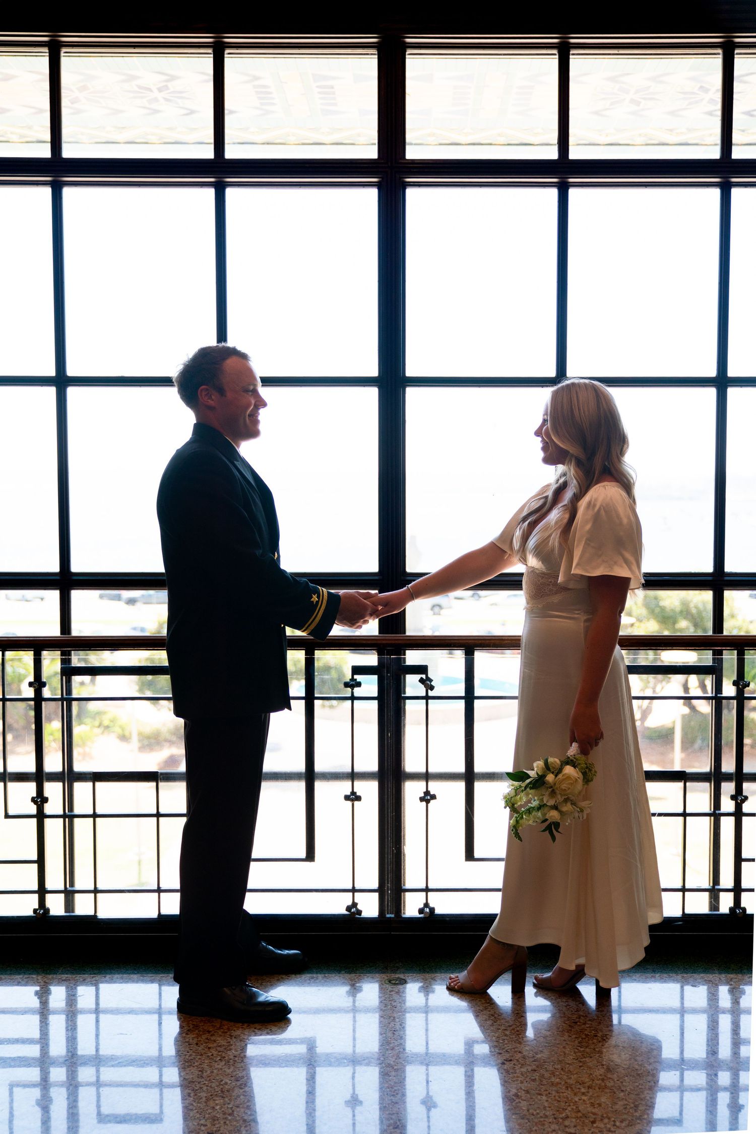 A couple stands in silhouette holding hands near large windows in an elegant indoor venue for a wedding ceremony.