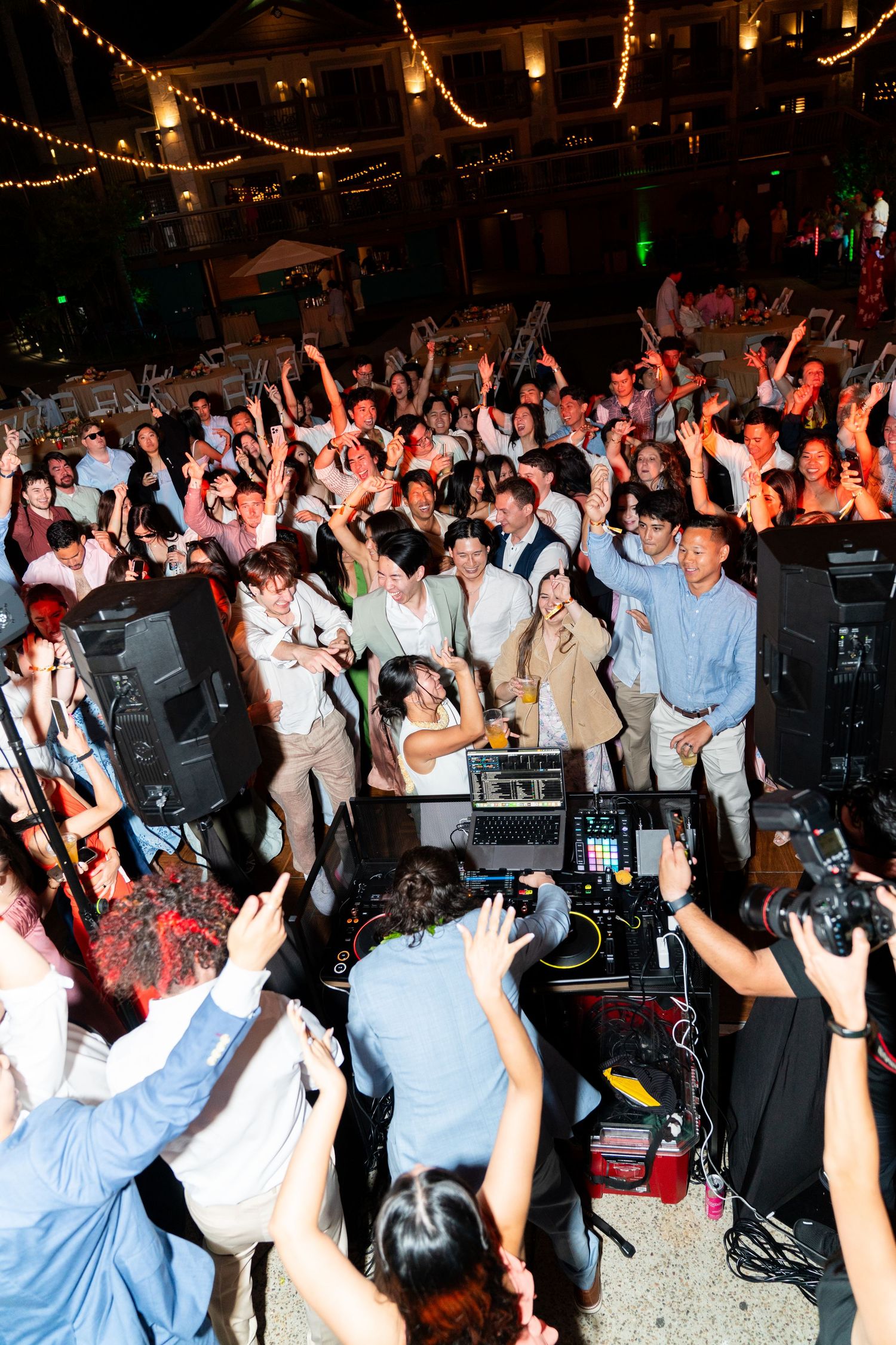 Energetic crowd with raised hands dancing at a nightclub around DJ equipment under string lights.