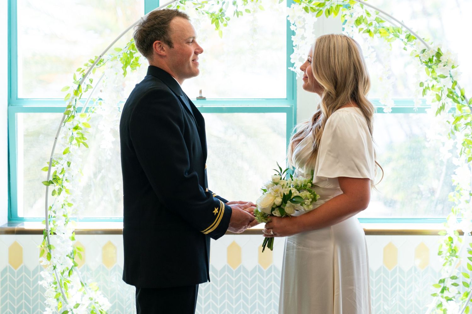 A couple stands together under an archway decorated with white flowers during an intimate wedding ceremony.