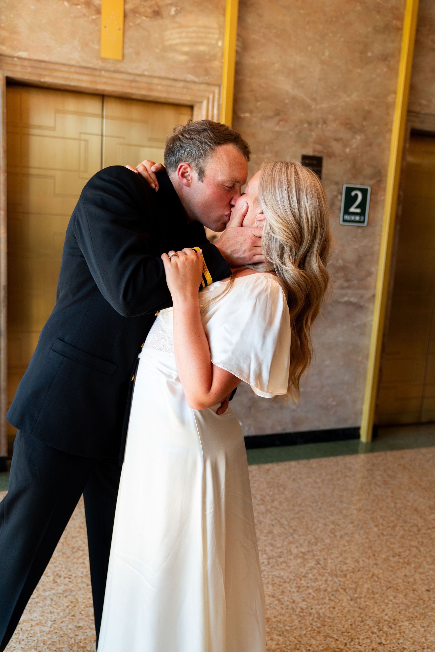 A couple shares a tender kiss in matching formal black and white attire against a marble hallway backdrop.