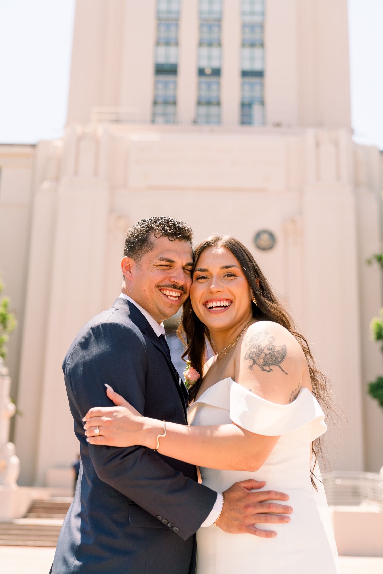 Happy couple embraces and laughs together in front of a tall building on their wedding day.