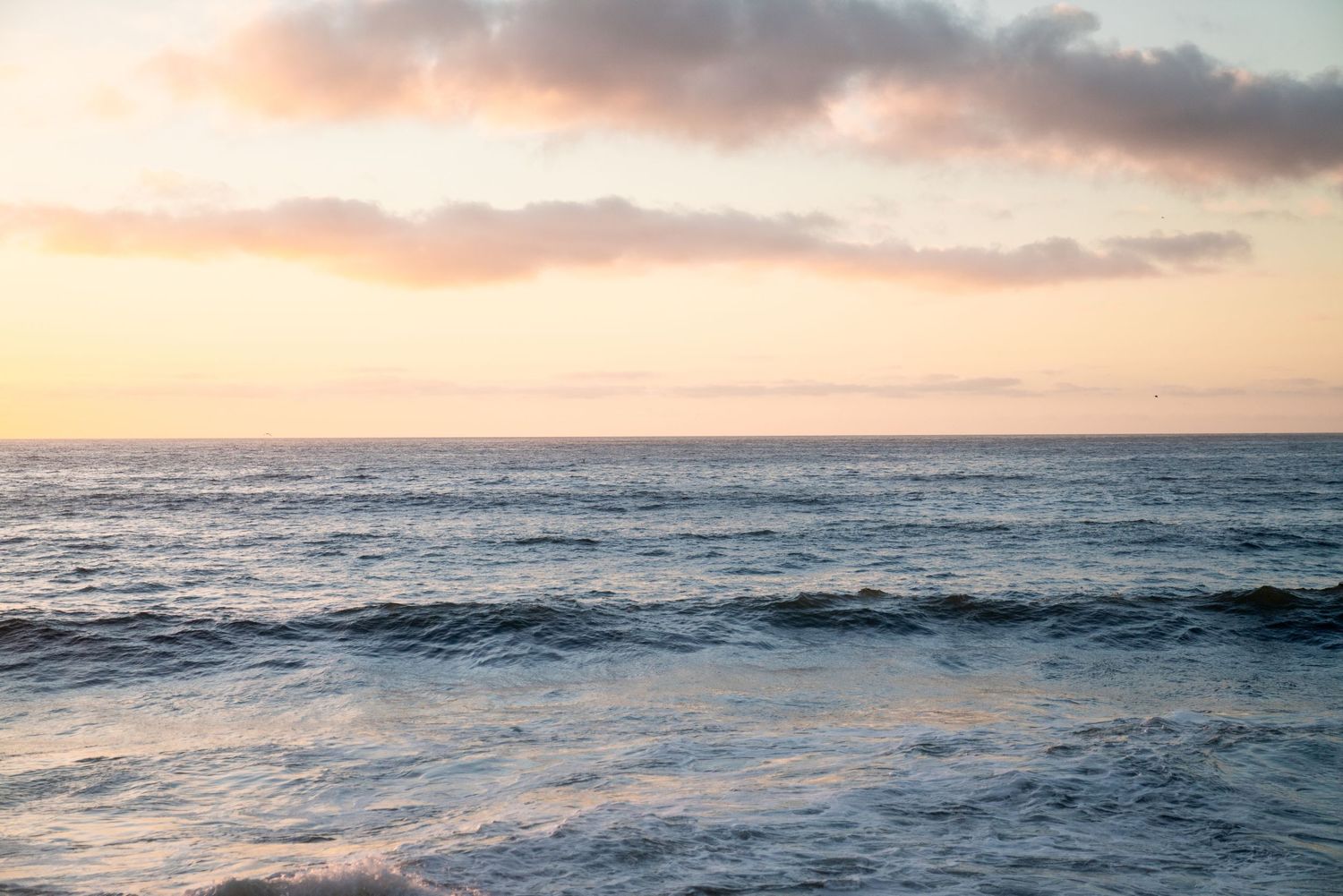 Soft ocean waves roll towards shore during a pastel pink and orange sunset over the peaceful horizon.