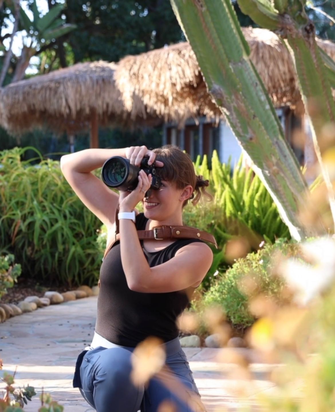 Photographer with a camera takes photos in a tropical garden setting with thatched roof structure in background.