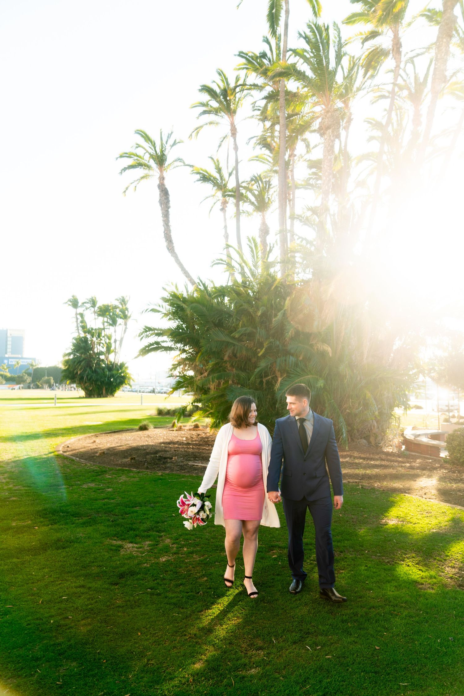A couple walks together on a grassy lawn with palm trees and golden sunlight streaming through the scene.