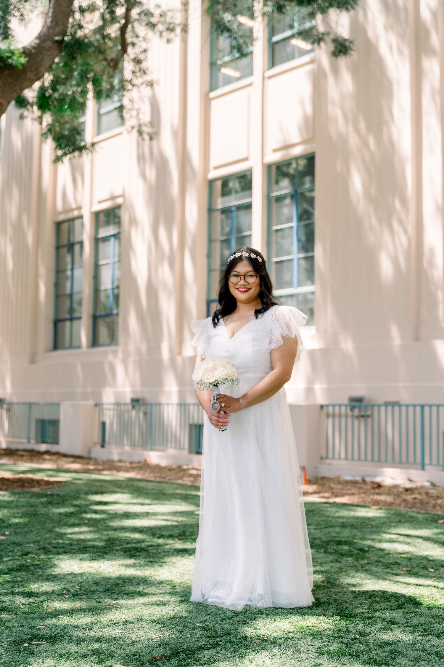 A bride in a flowing white ball gown poses gracefully on a sunny lawn in front of a building with tall windows.