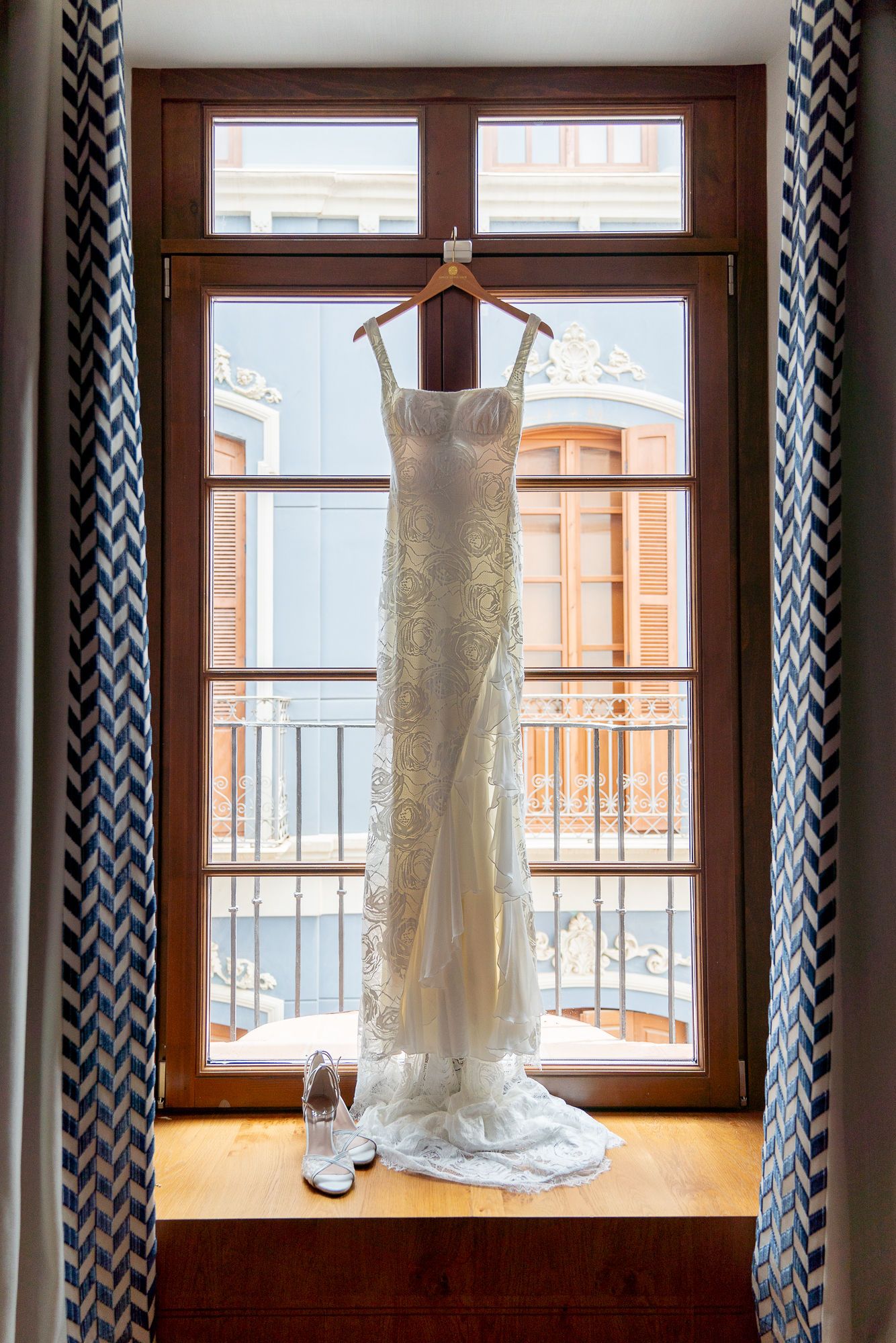 A white lace wedding dress hangs elegantly in a wooden window frame, with white bridal shoes on the windowsill.