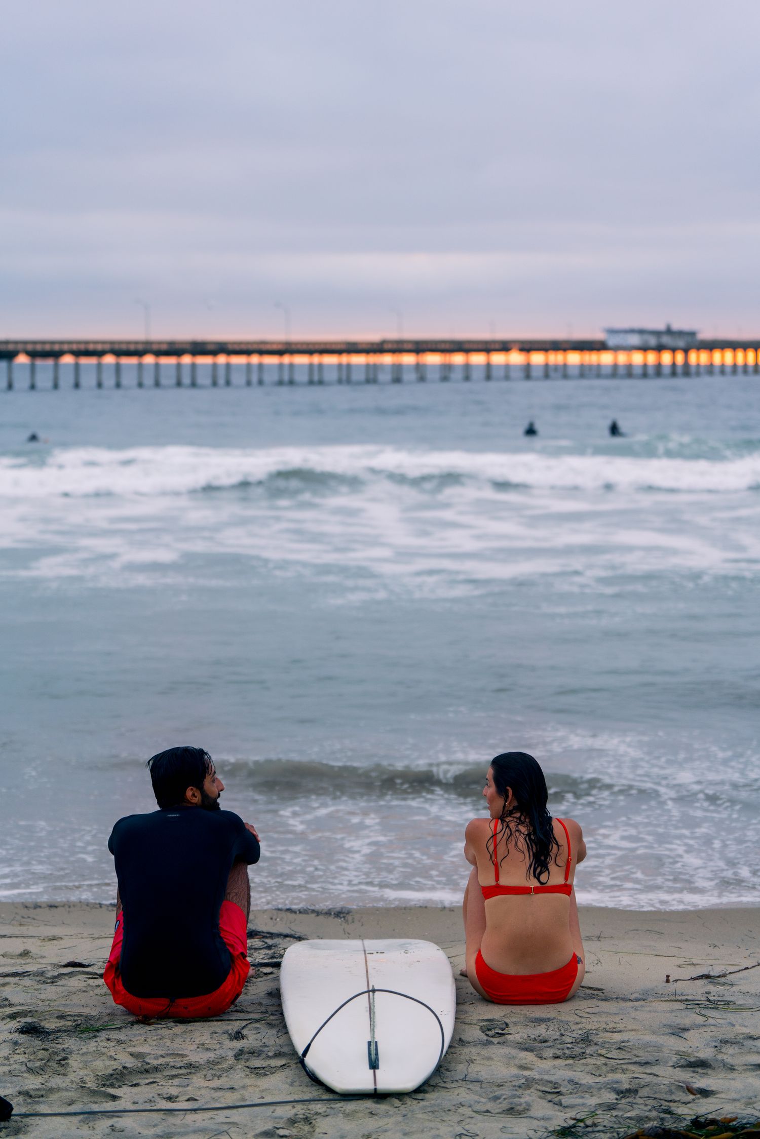Two surfers sit on the beach with a surfboard watching waves roll in at sunset near a long pier over the ocean.