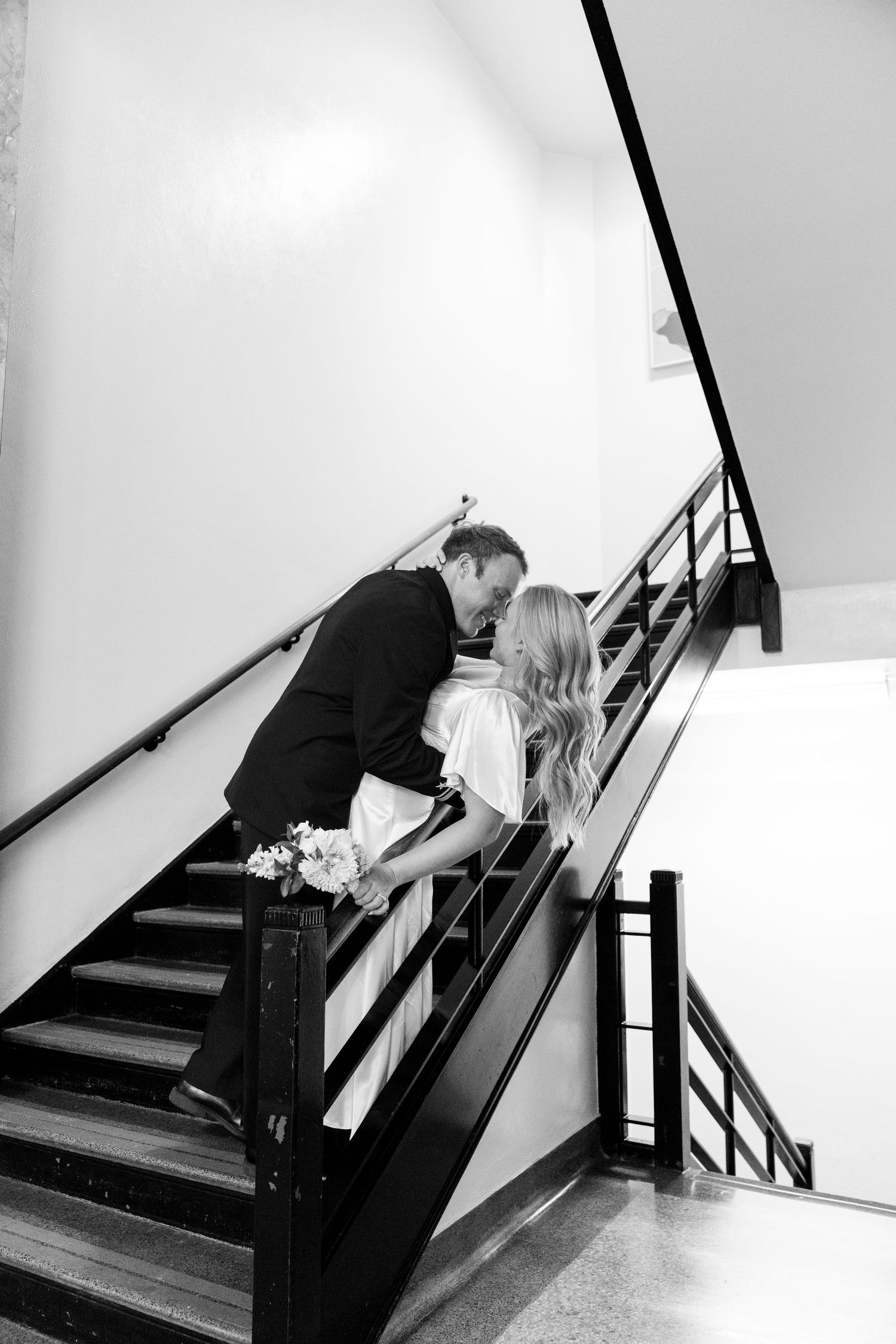 A romantic black and white photo of a couple sharing a kiss on a staircase with black railings.