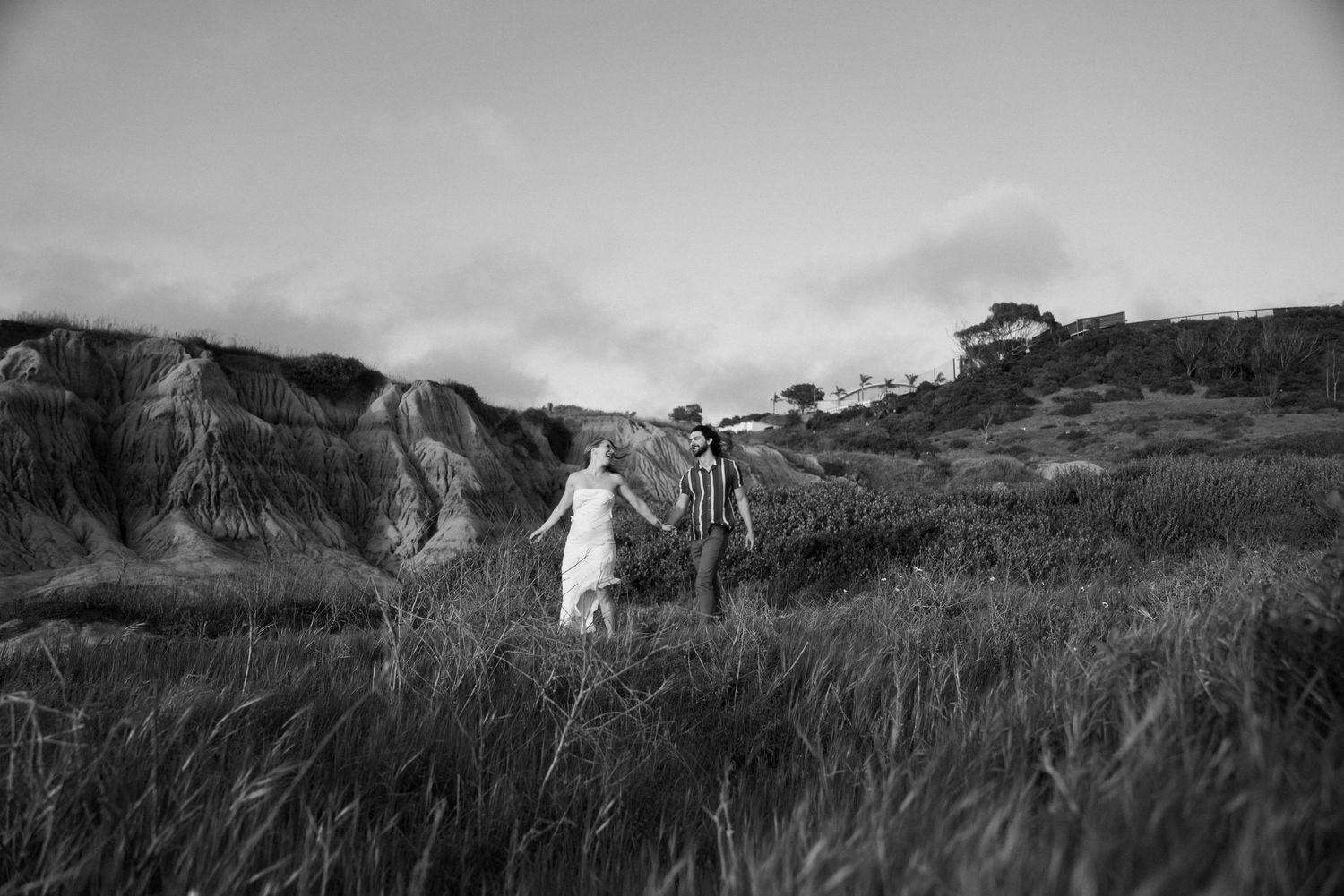 A graceful figure in white walks through tall windswept grass against a backdrop of rugged coastal cliffs in black and white.