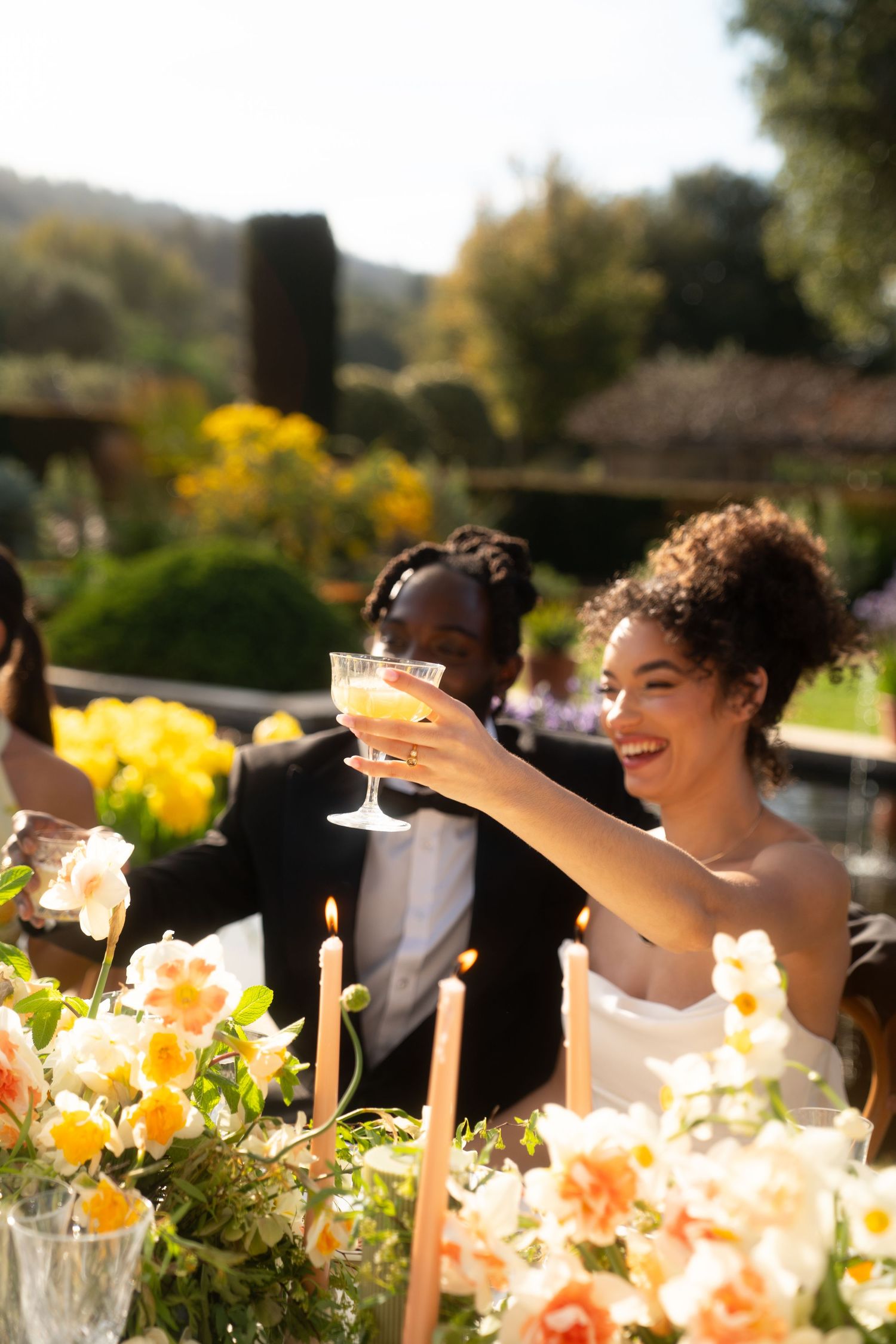Wedding guests enjoying cocktails and champagne at an outdoor garden reception with peach and yellow floral decorations.
