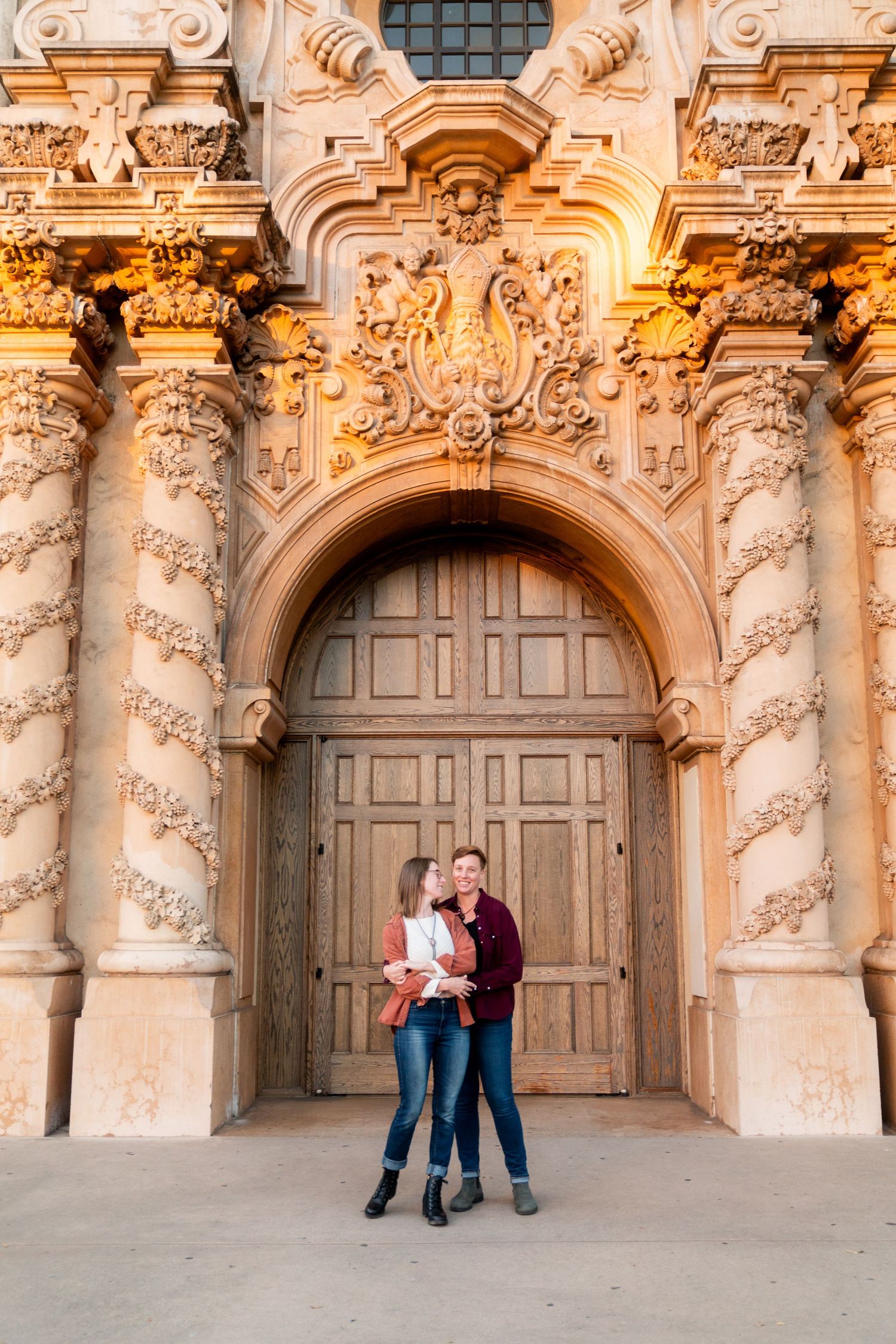 A couple stands in front of an ornate Baroque doorway with spiral columns and intricate stone carvings at sunset.