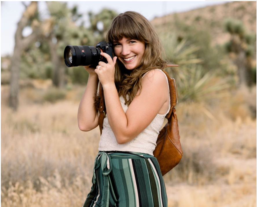 Professional photographer in flowing dress captures outdoor shots in desert landscape with tall grass and shrubs.